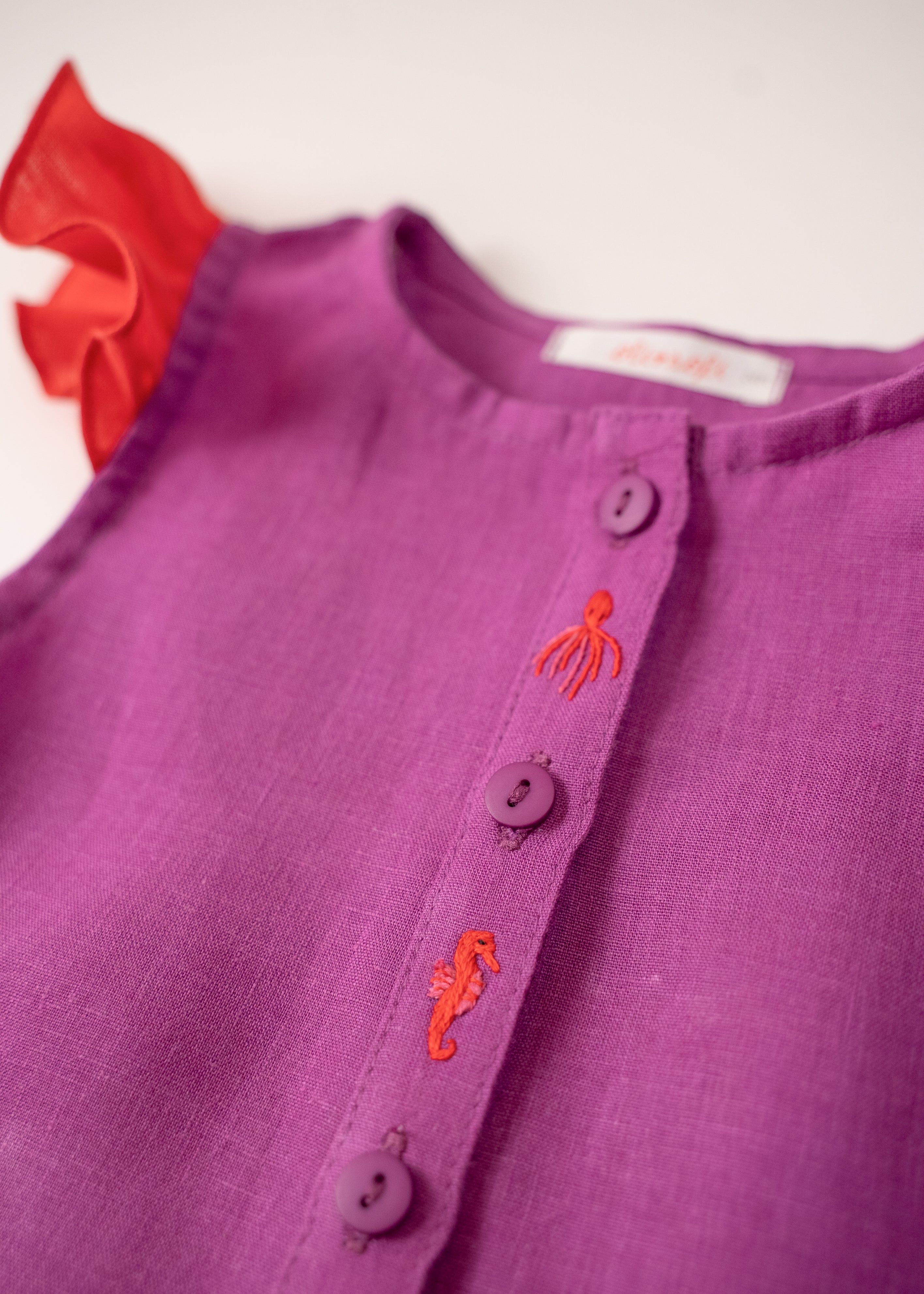 Detalhe dos bordados à mão de polvo e cavalo marinho da camisa infantil menina em linho roxo com mangas de babado vermelho vibrante.