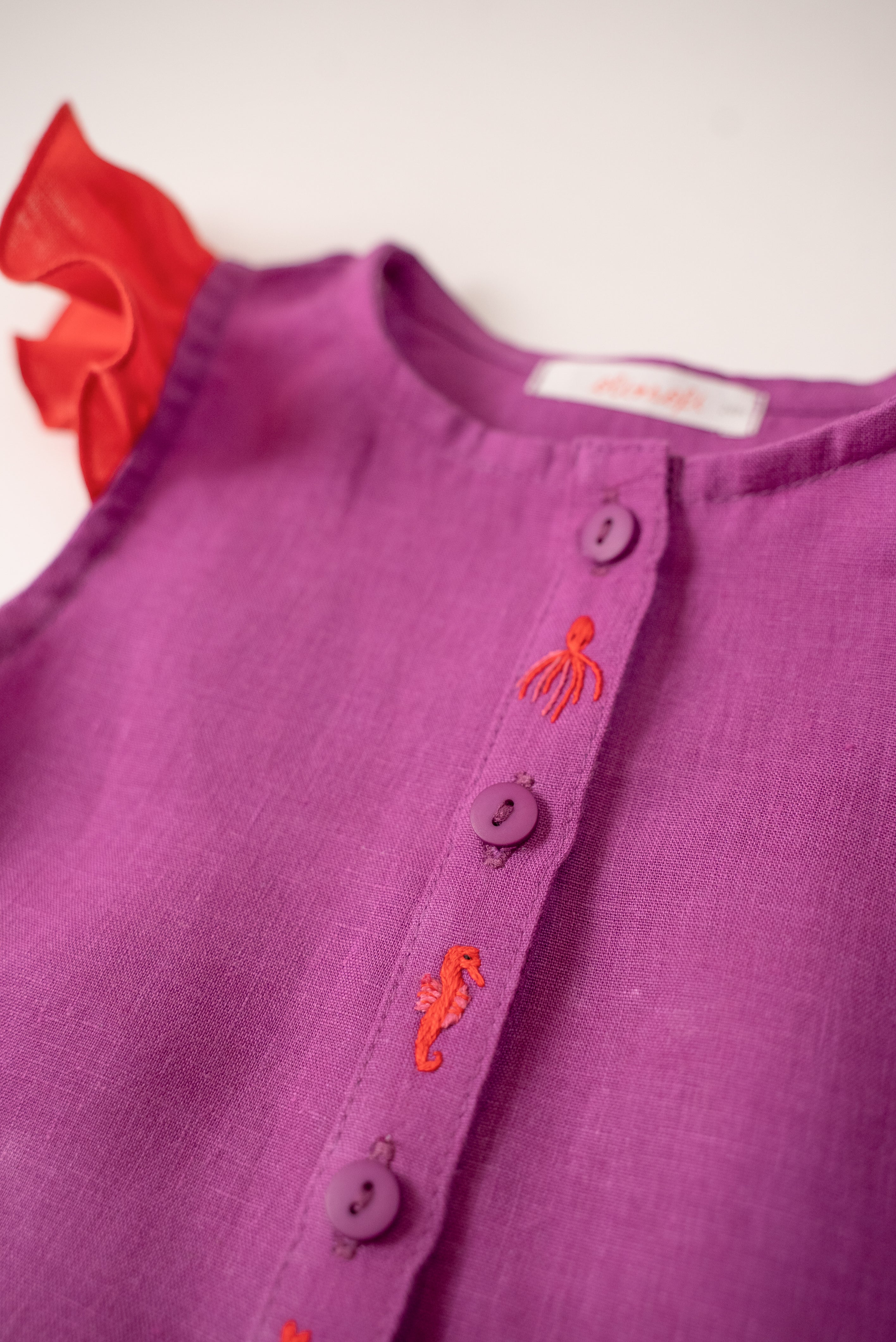 Detalhe dos bordados à mão de polvo e cavalo marinho da camisa infantil menina em linho roxo com mangas de babado vermelho vibrante.