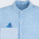 Camisa infantil em linho azul com bolso listrado e tubarão bordado à mão, vista de frente em detalhe.