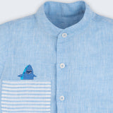 Camisa infantil em linho azul com bolso listrado e tubarão bordado à mão, vista de frente em detalhe.