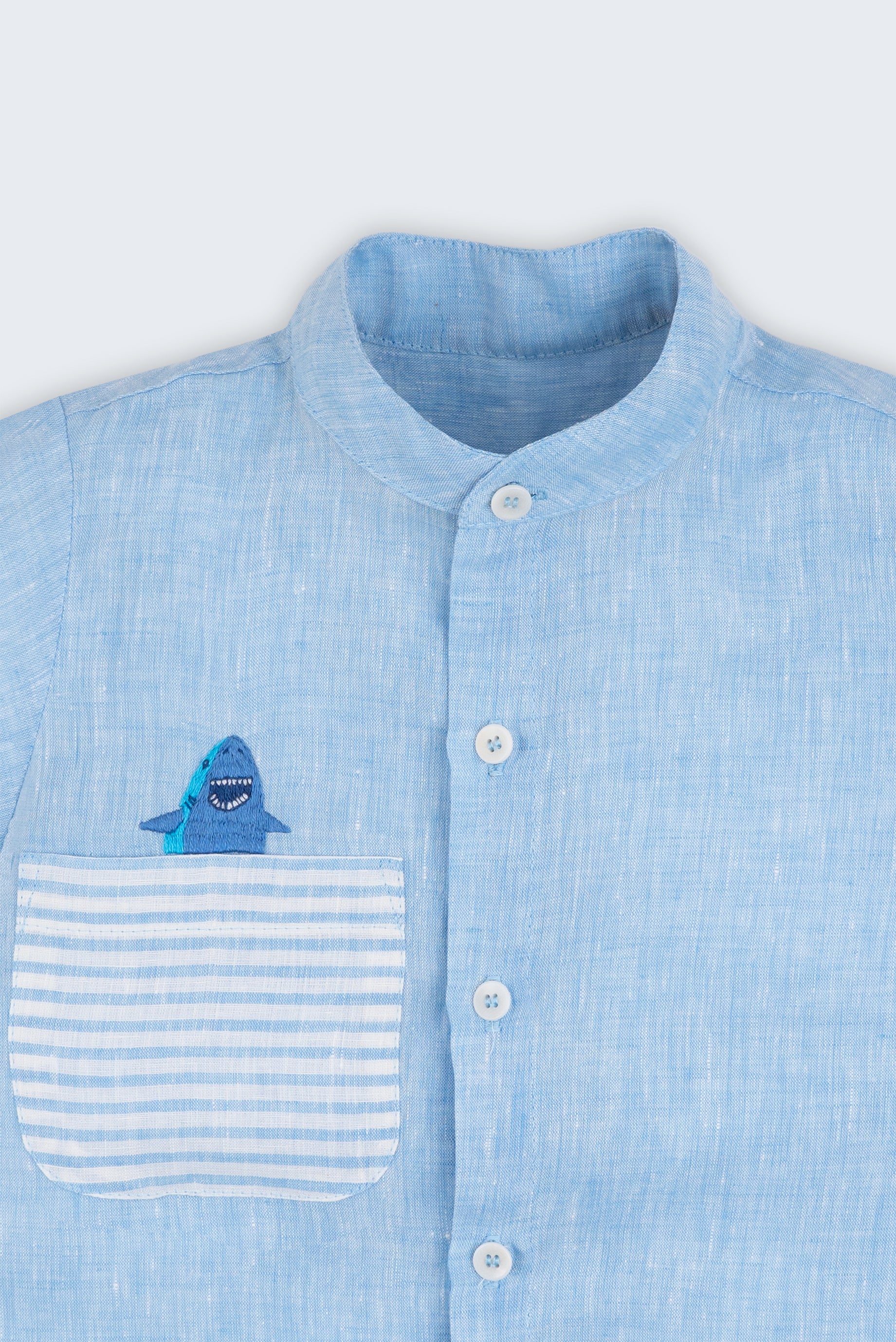 Camisa infantil em linho azul com bolso listrado e tubarão bordado à mão, vista de frente em detalhe.