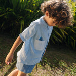 Menino olhando para baixo usando camisa infantil de linho azul com bolso listrado e tubarão bordado à mão.