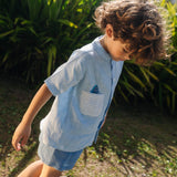 Menino olhando para baixo usando camisa infantil de linho azul com bolso listrado e tubarão bordado à mão.
