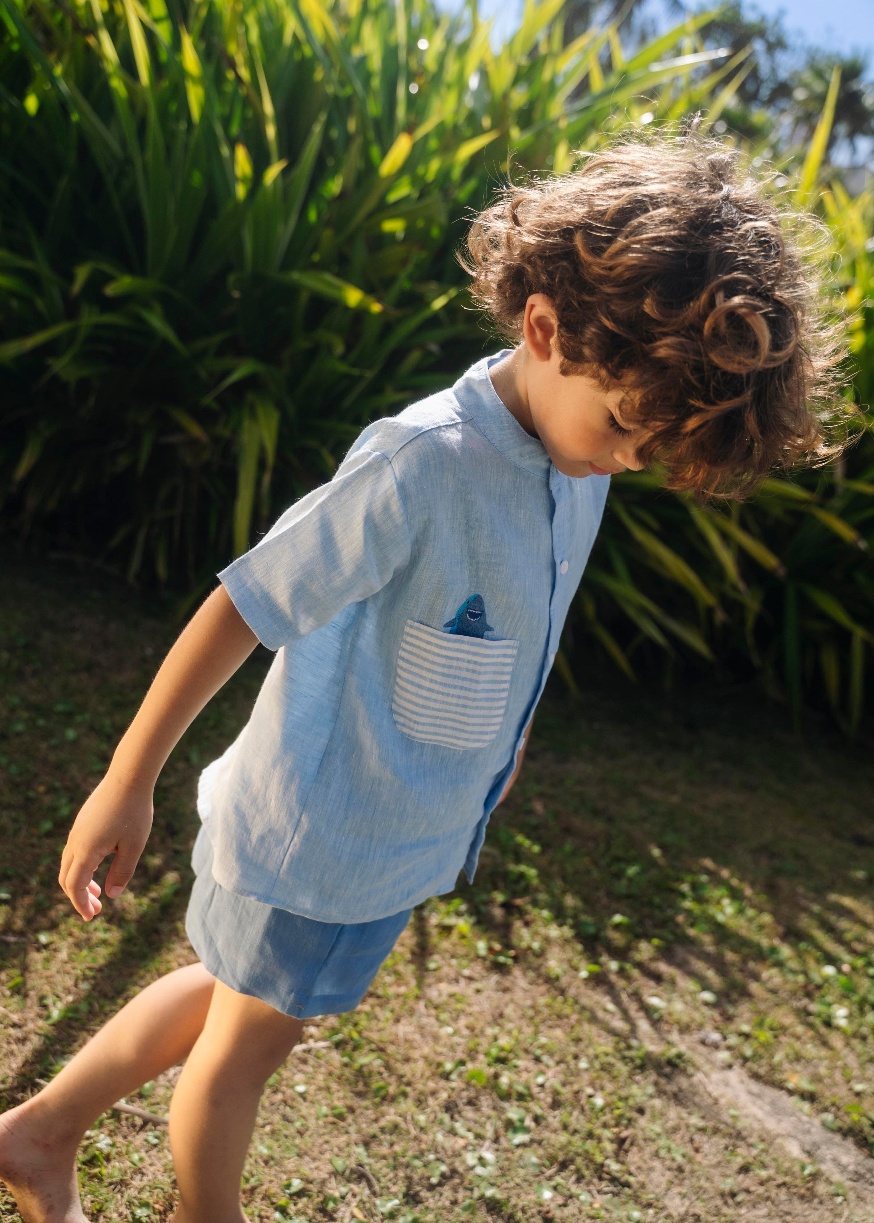 Menino olhando para baixo usando camisa infantil de linho azul com bolso listrado e tubarão bordado à mão.
