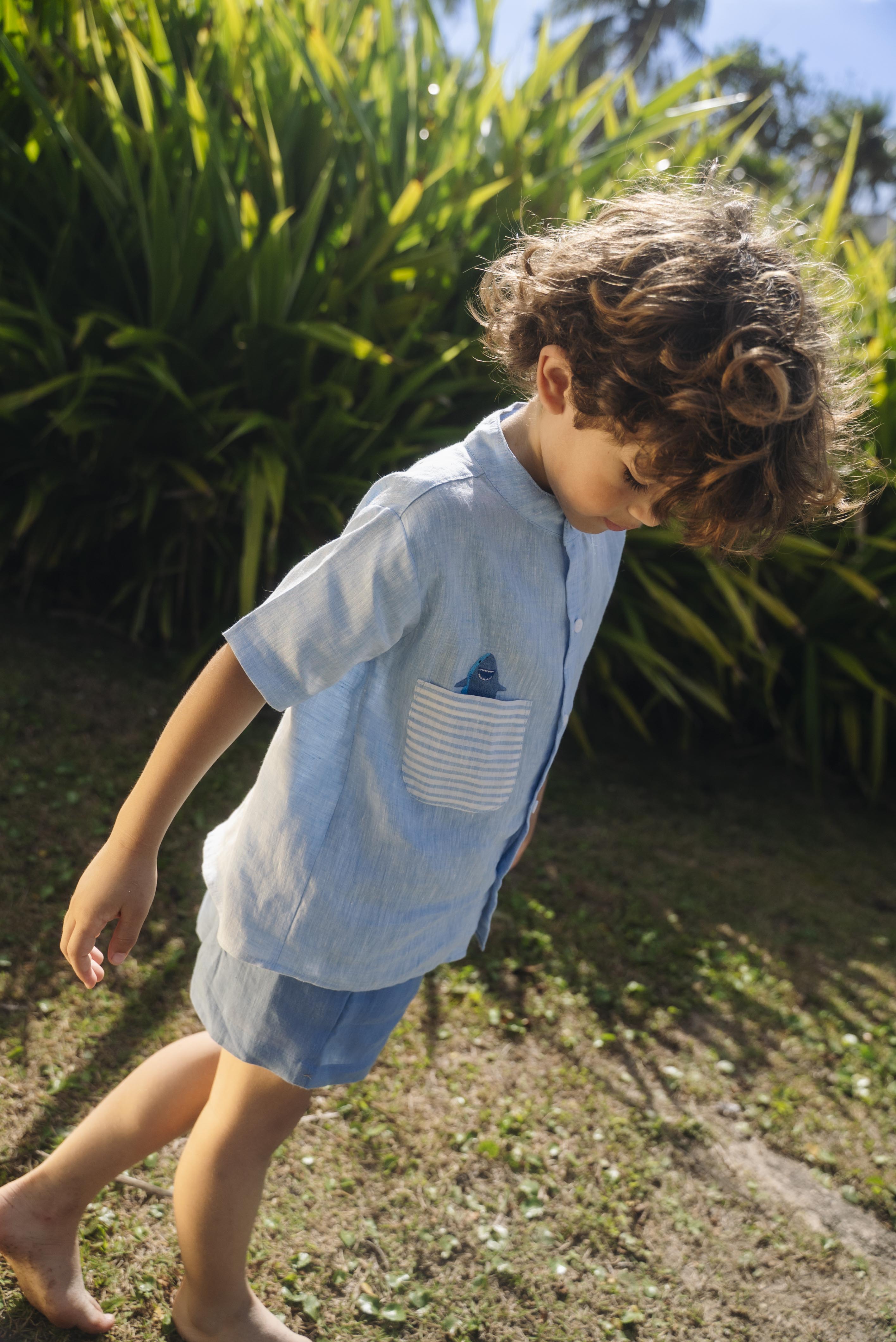Menino olhando para baixo usando camisa infantil de linho azul com bolso listrado e tubarão bordado à mão.