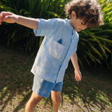 Menino usando camisa infantil de linho azul com bolso listrado e tubarão bordado à mão.
