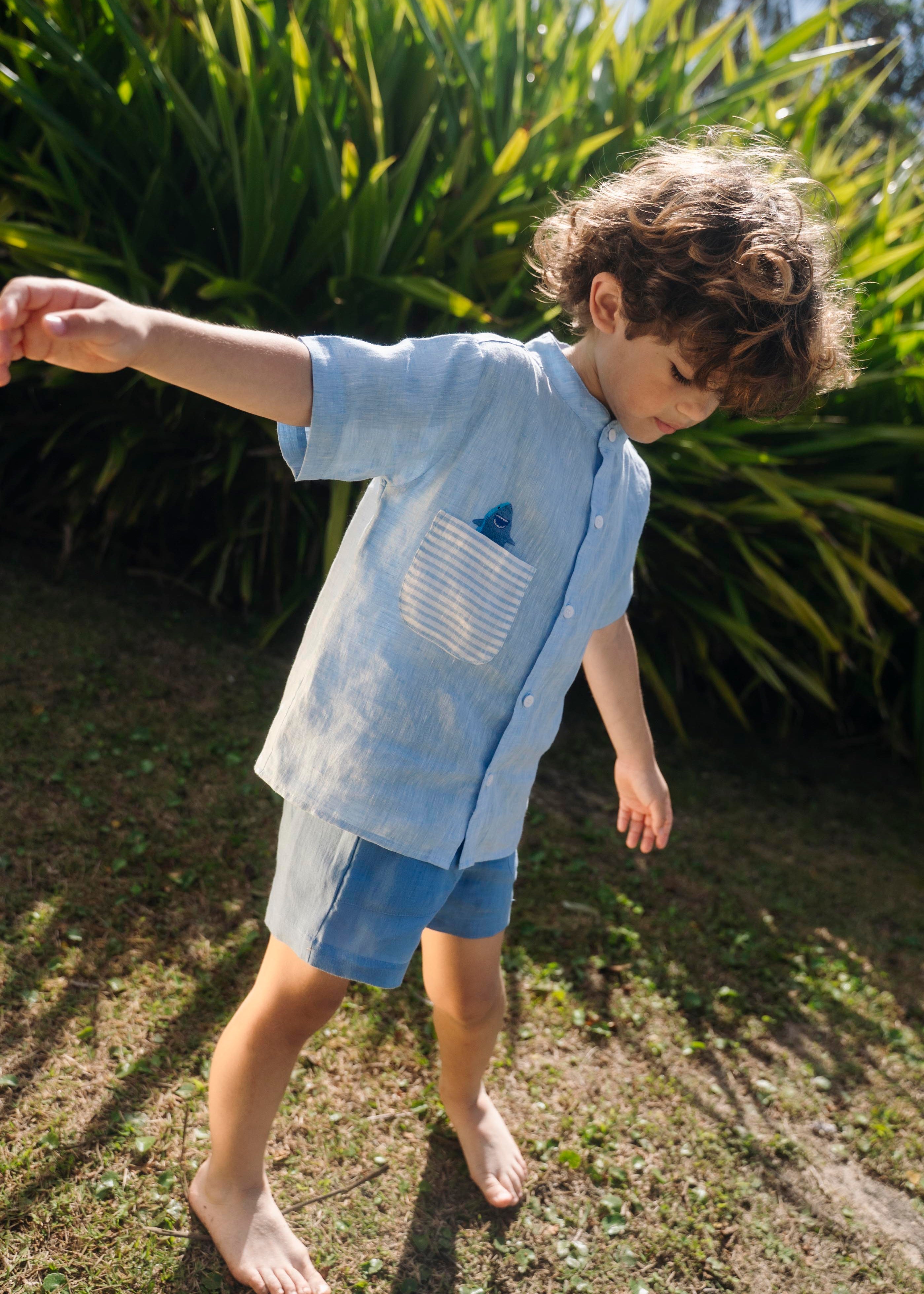 Menino usando camisa infantil de linho azul com bolso listrado e tubarão bordado à mão.