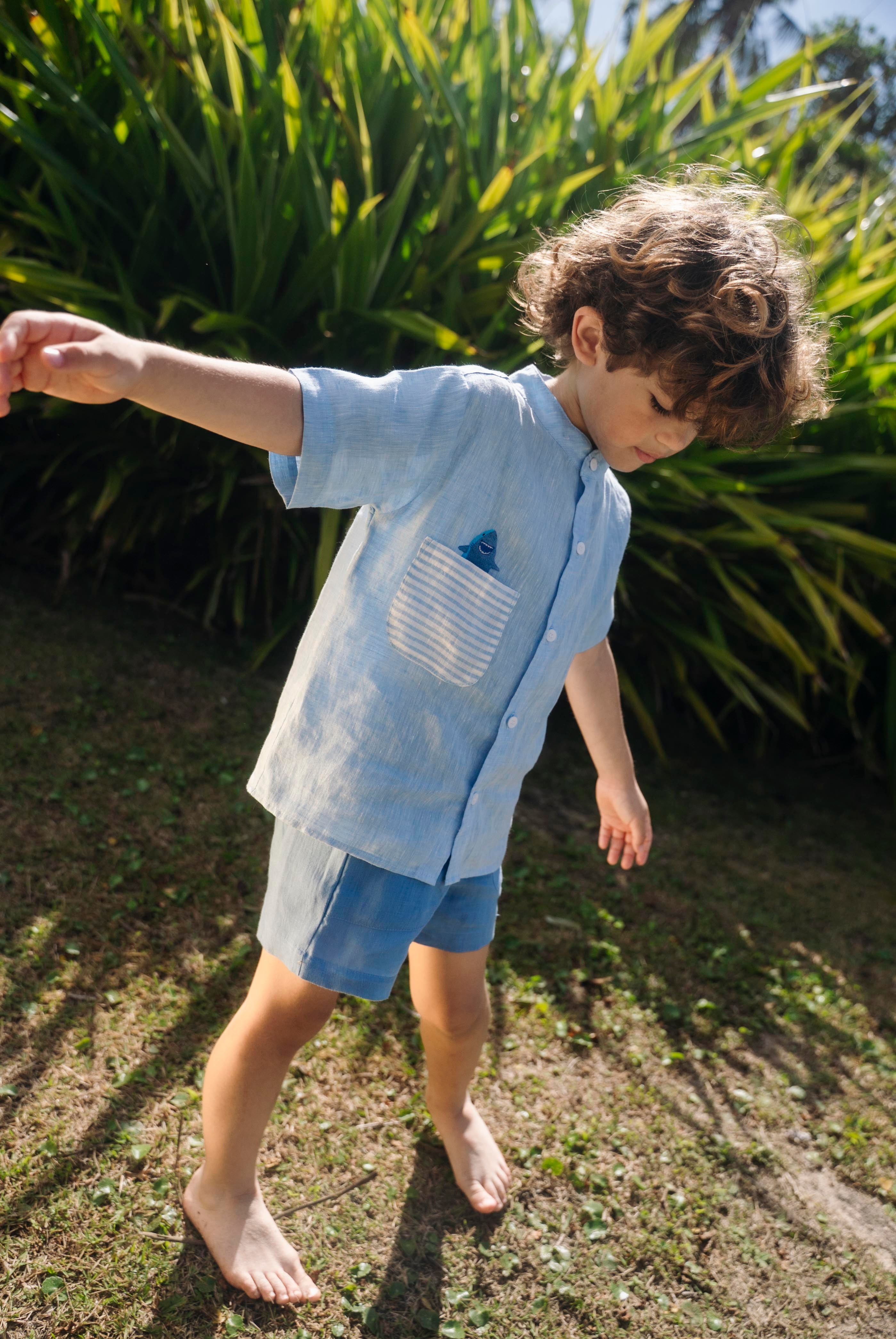 Menino usando camisa infantil de linho azul com bolso listrado e tubarão bordado à mão.