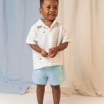 Menino em pé usando camisa infantil de linho listrada com bordados de patinhos e shorts azul claro