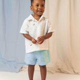 Menino em pé usando camisa infantil de linho listrada com bordados de patinhos e shorts azul claro