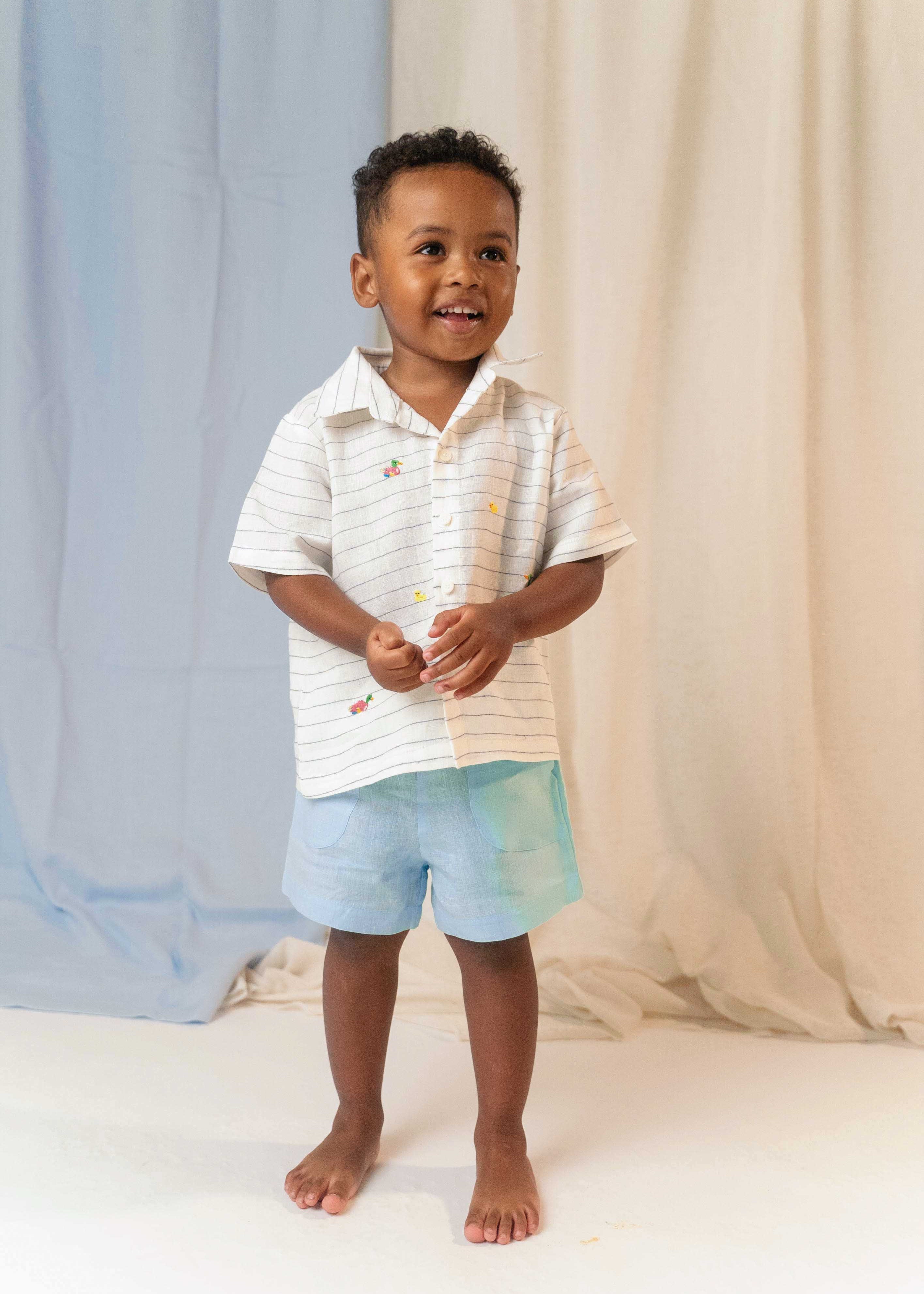 Menino em pé usando camisa infantil de linho listrada com bordados de patinhos e shorts azul claro
