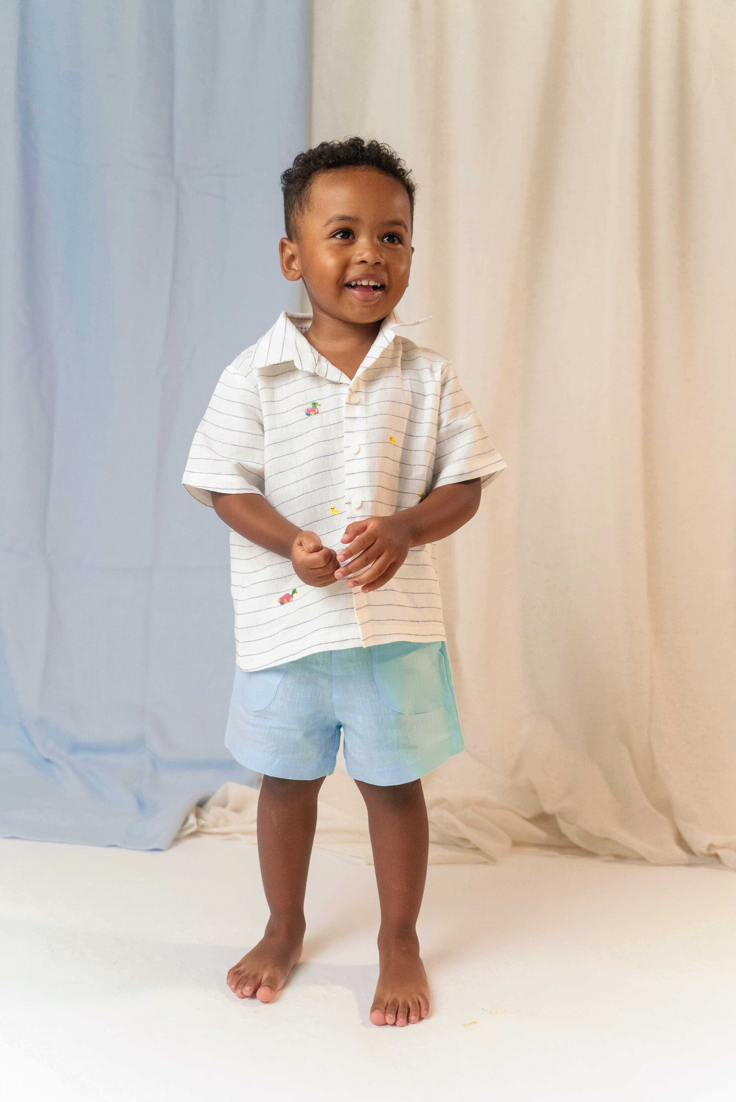 Menino em pé usando camisa infantil de linho listrada com bordados de patinhos e shorts azul claro