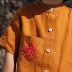 Close de camisa em linho terracota terracota com polvo bordado no bolso e botões brancos contrastando.
