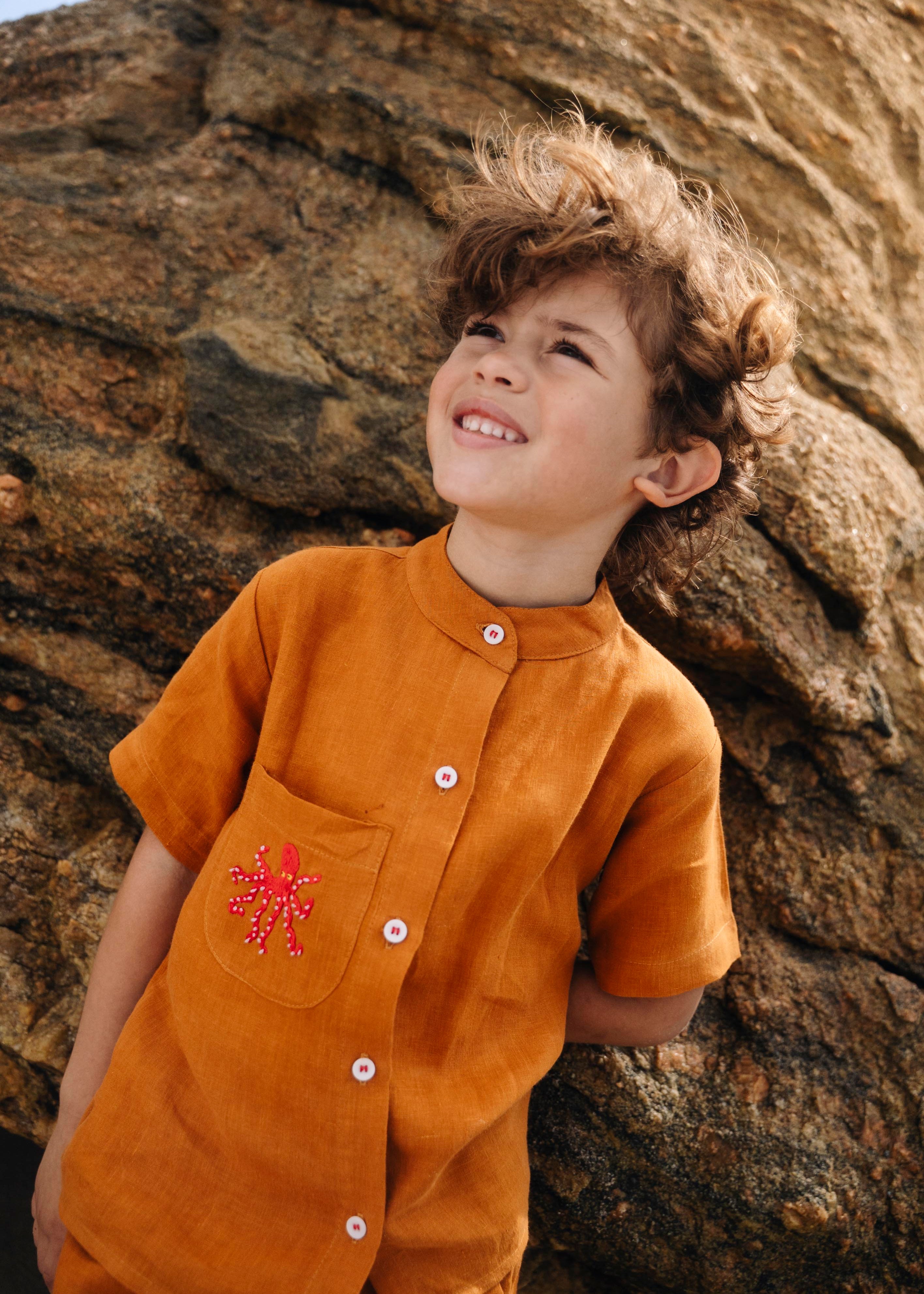 Close de menino com camisa em linho terracota com polvo bordado no bolso e botões brancos contrastando.