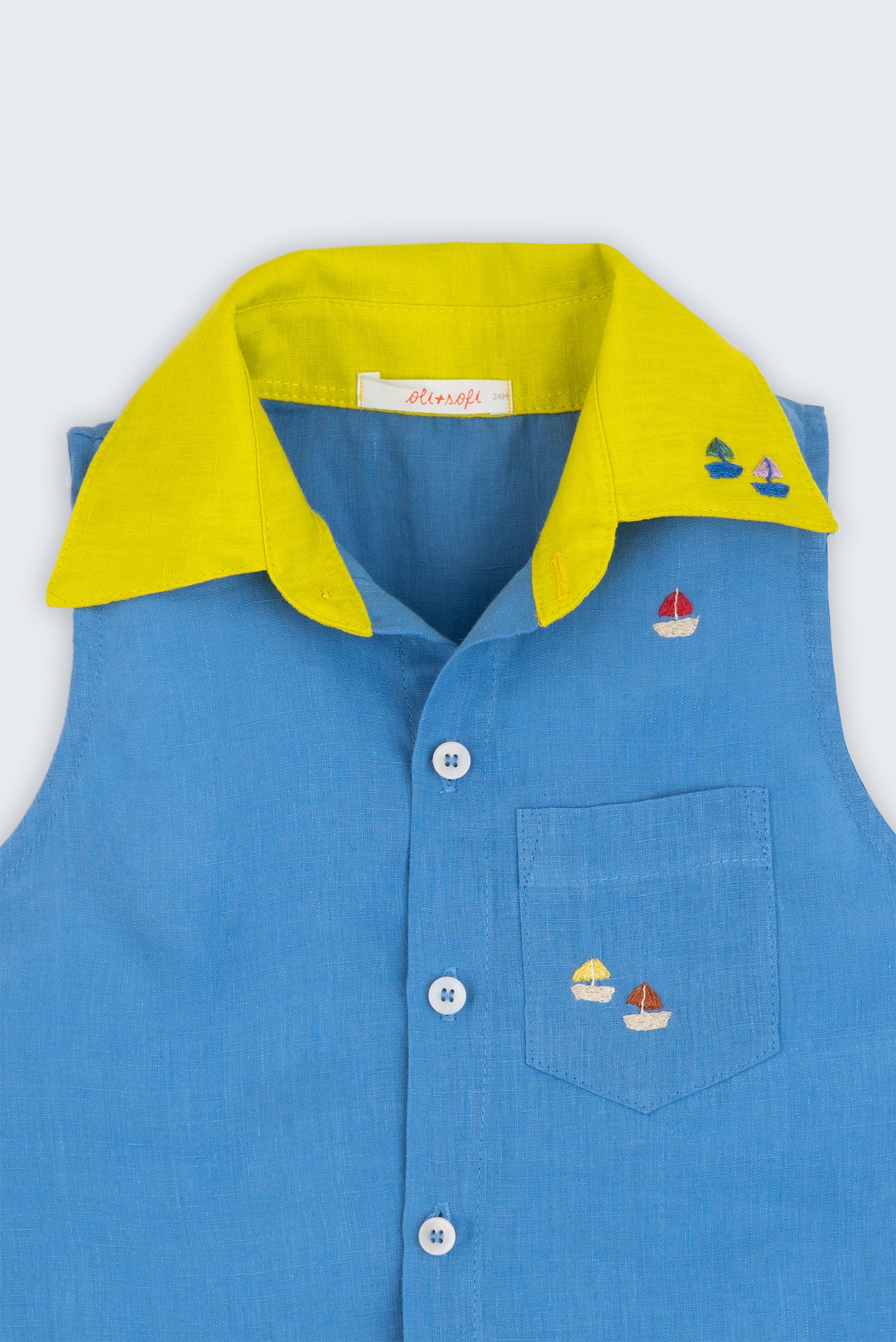 Camisa infantil em linho azul com gola amarelo lima e bordado de barquinhos coloridos espalhados pela pala, ombro e bolso feito à mão, vista de frente em detalhe.