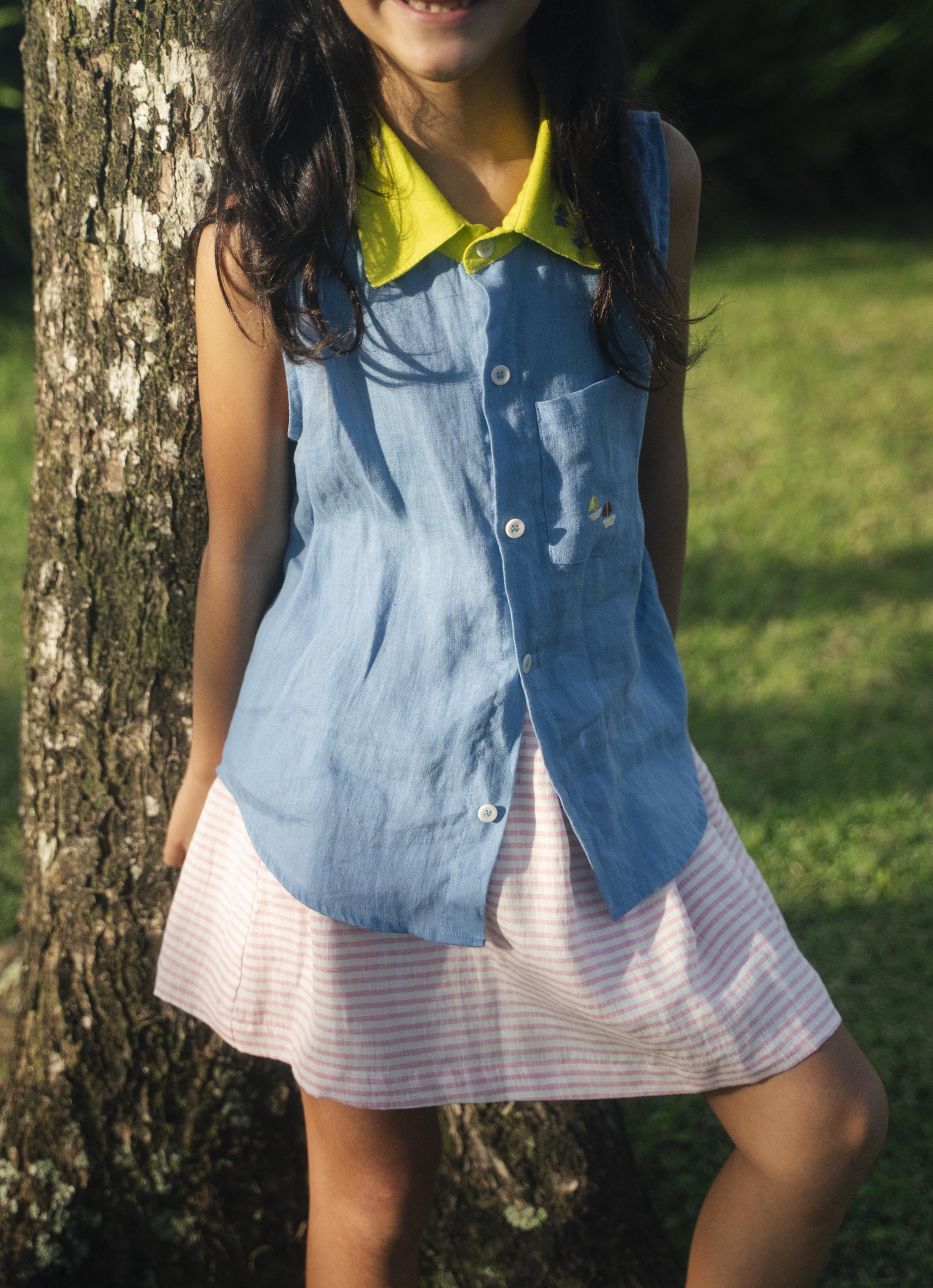 Menina com camisa infantil em linho azul com gola amarelo lima e bordados de barquinhos coloridos com saia listrada rosa e branco.
