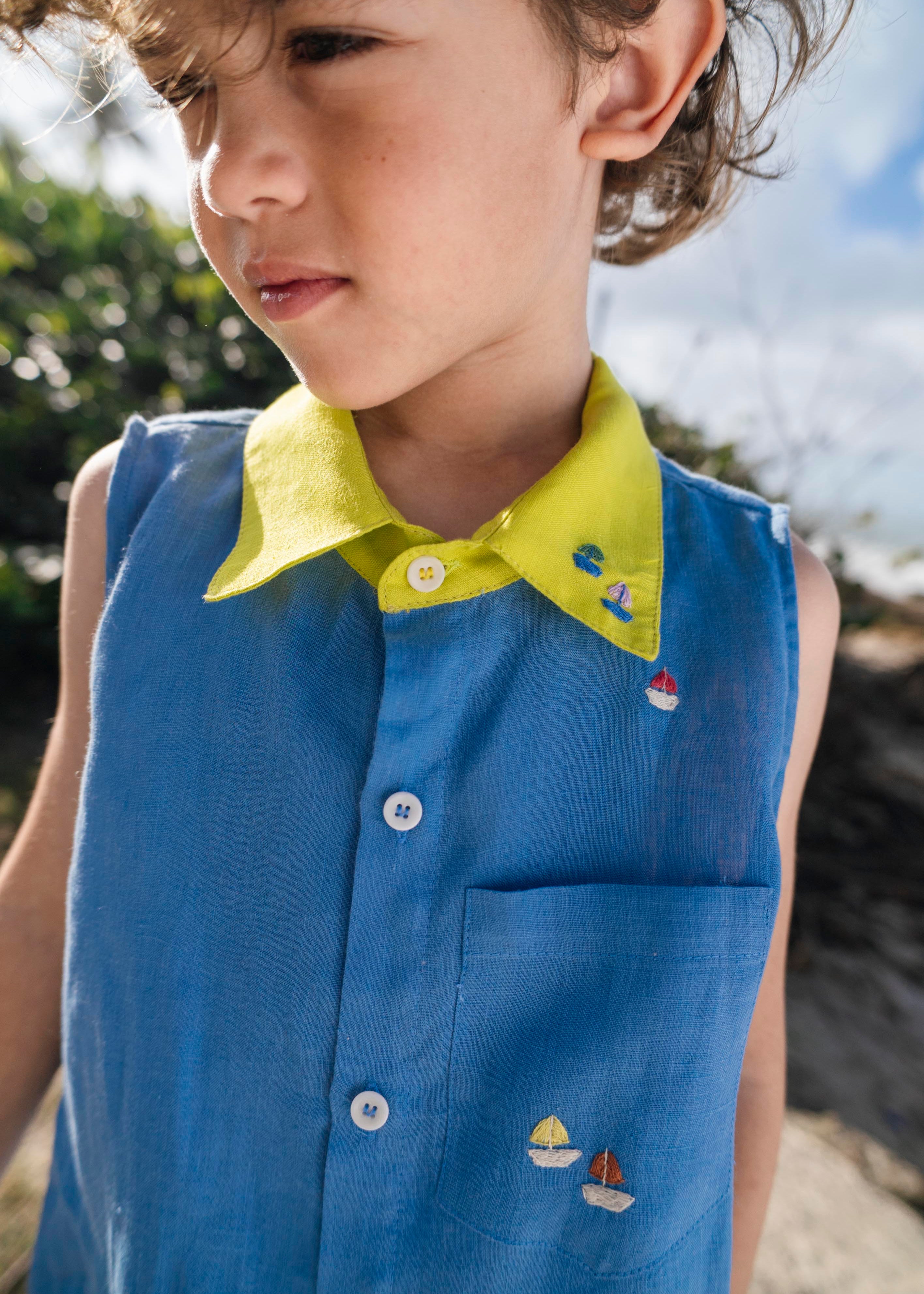 Menino usando camisa infantil em linho azul com gola amarelo lima e bordados de barquinhos coloridos espalhados pela pala, ombro e bolso feitos à mão.