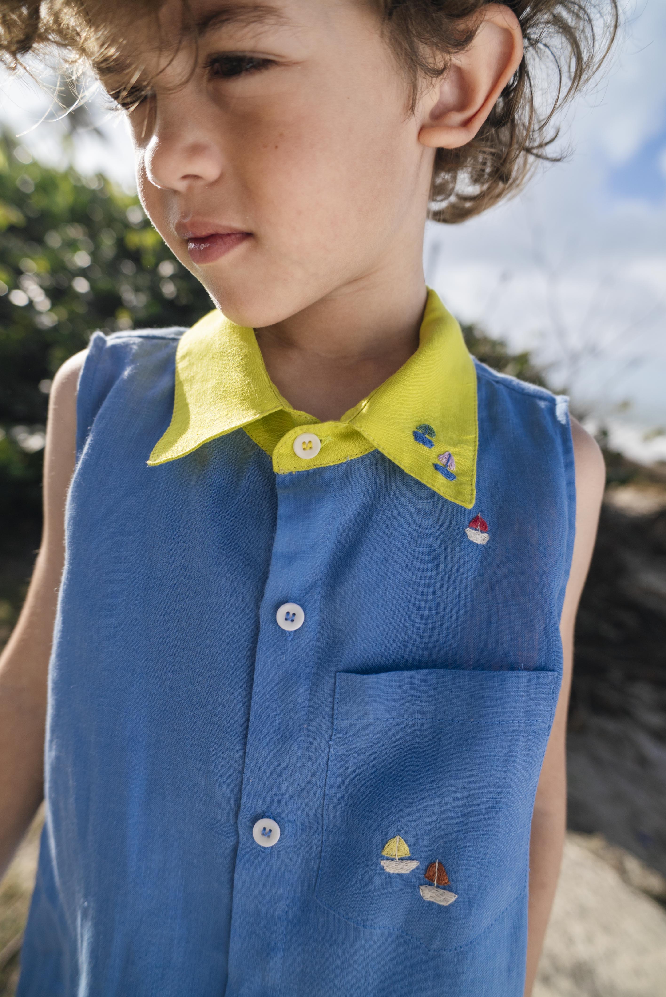 Menino usando camisa infantil em linho azul com gola amarelo lima e bordados de barquinhos coloridos espalhados pela pala, ombro e bolso feitos à mão.