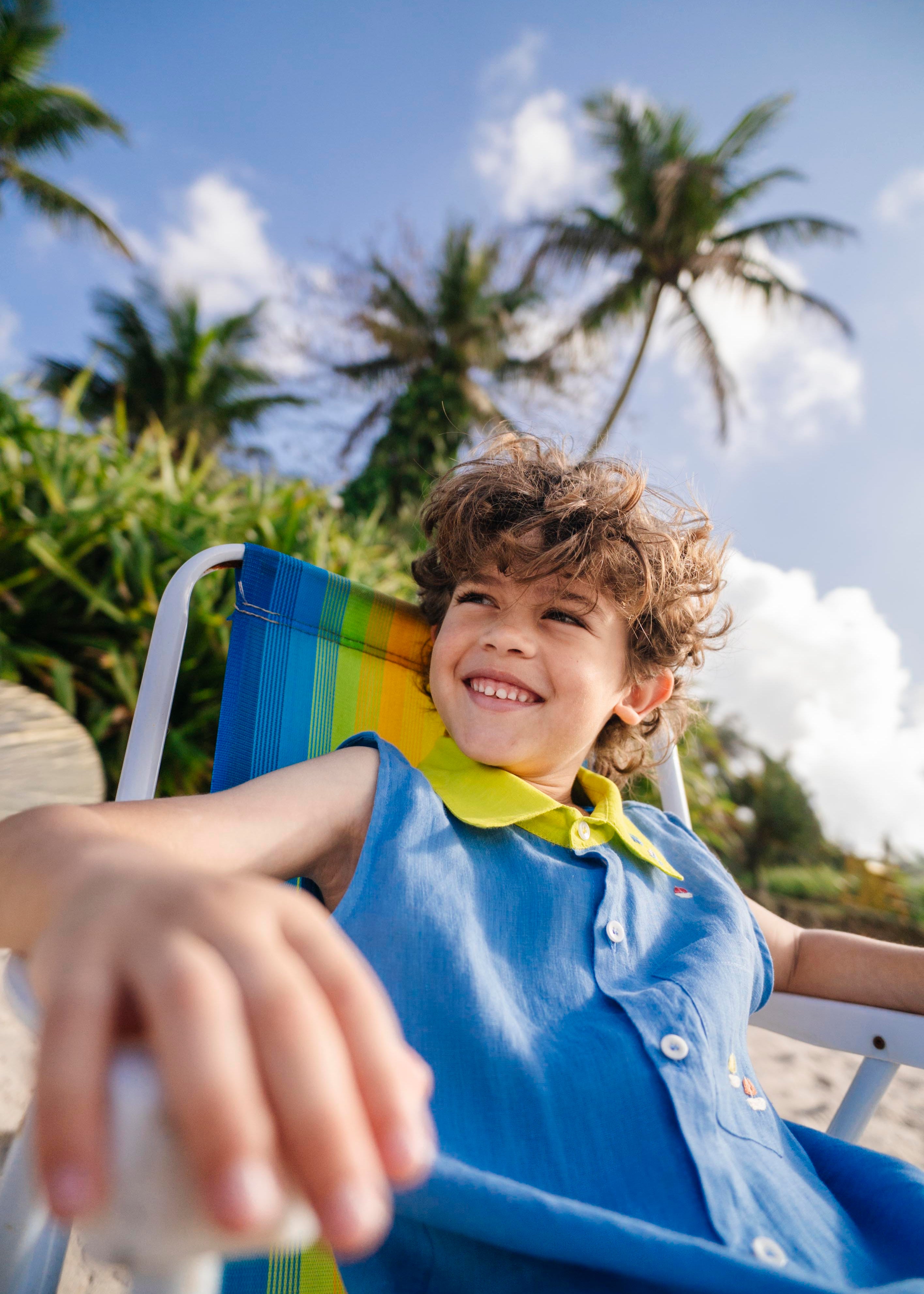 Menino sentado em cadeira de praia, com camisa em linho azul com gola amarelo lima e bordado de barquinhos.