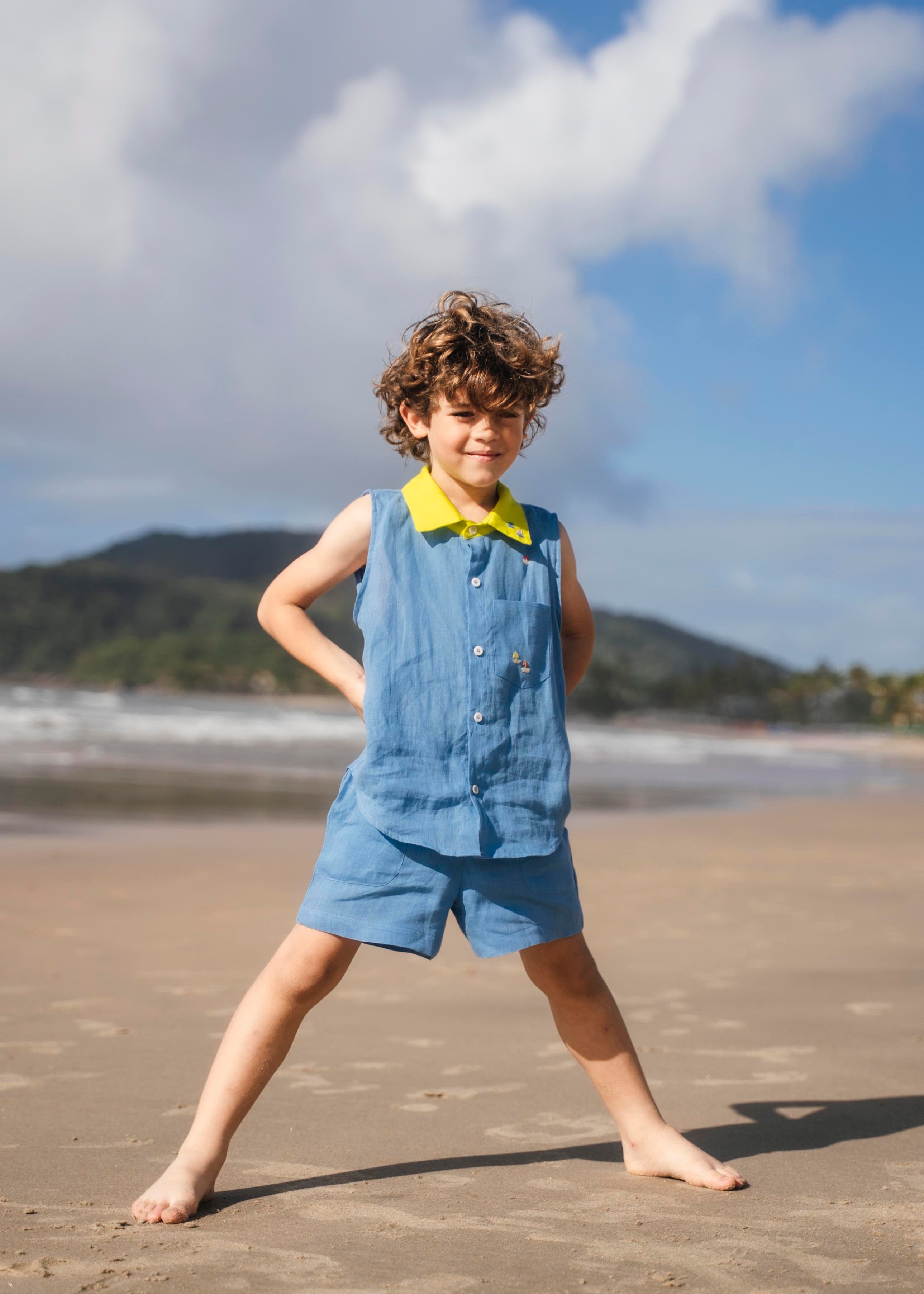 Menino usando camisa infantil em linho azul com gola amarelo lima e bordado de barquinhos coloridos feitos à mão, combinando com shorts em mesmo tom.