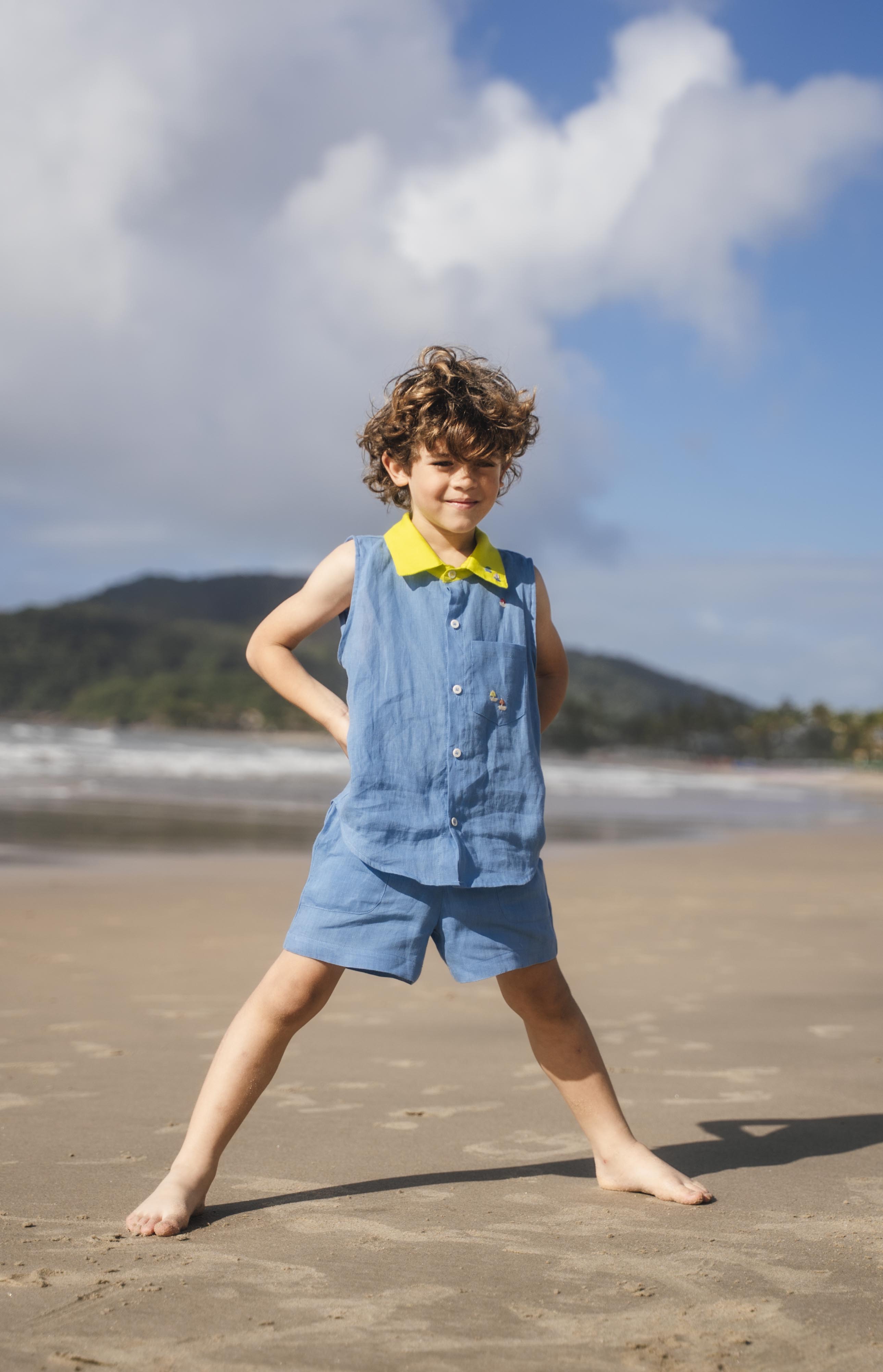 Menino usando camisa infantil em linho azul com gola amarelo lima e bordado de barquinhos coloridos feitos à mão, combinando com shorts em mesmo tom.