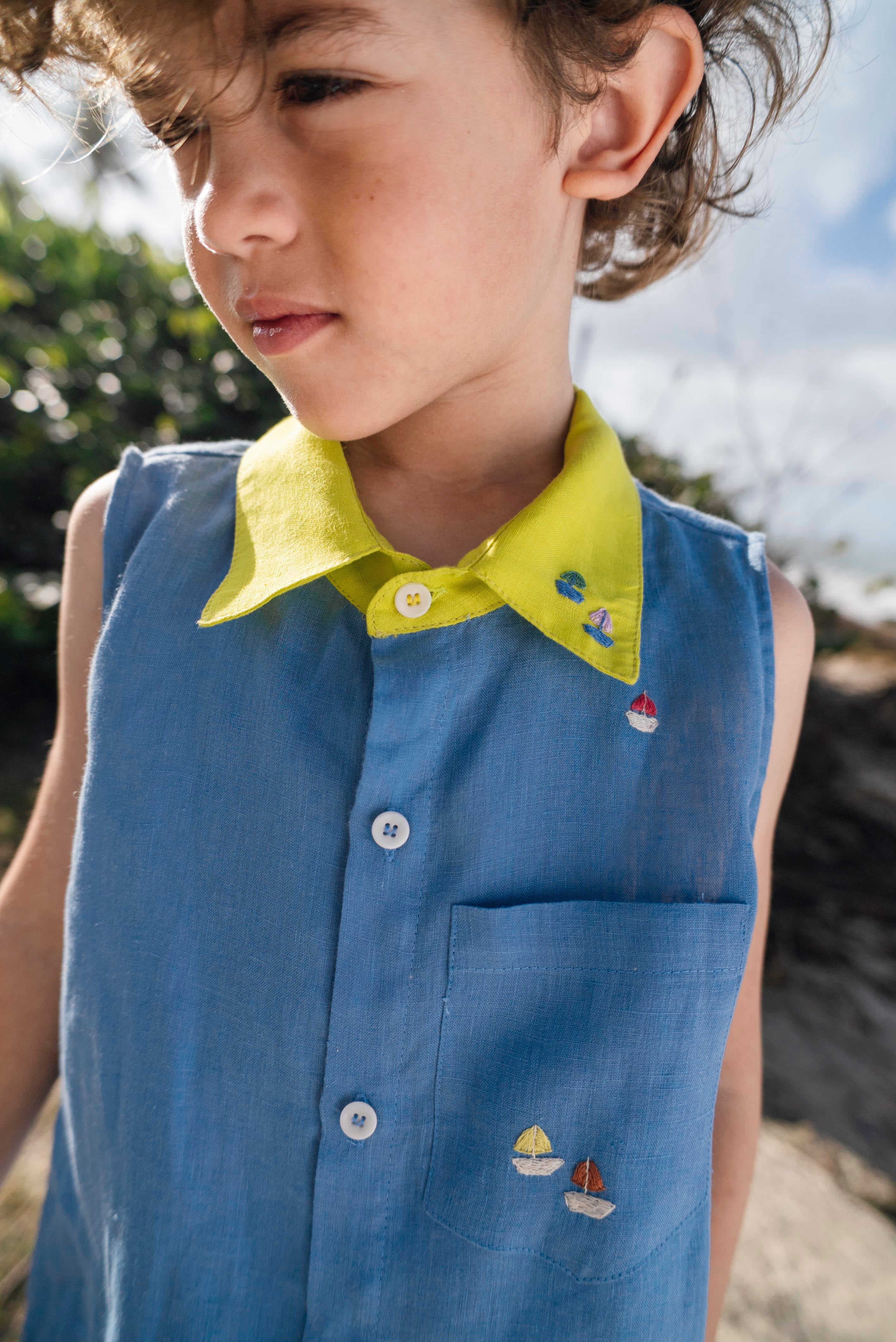 Menino usando camisa infantil em linho azul com gola amarelo lima e bordados de barquinhos coloridos espalhados pela pala, ombro e bolso feitos à mão.