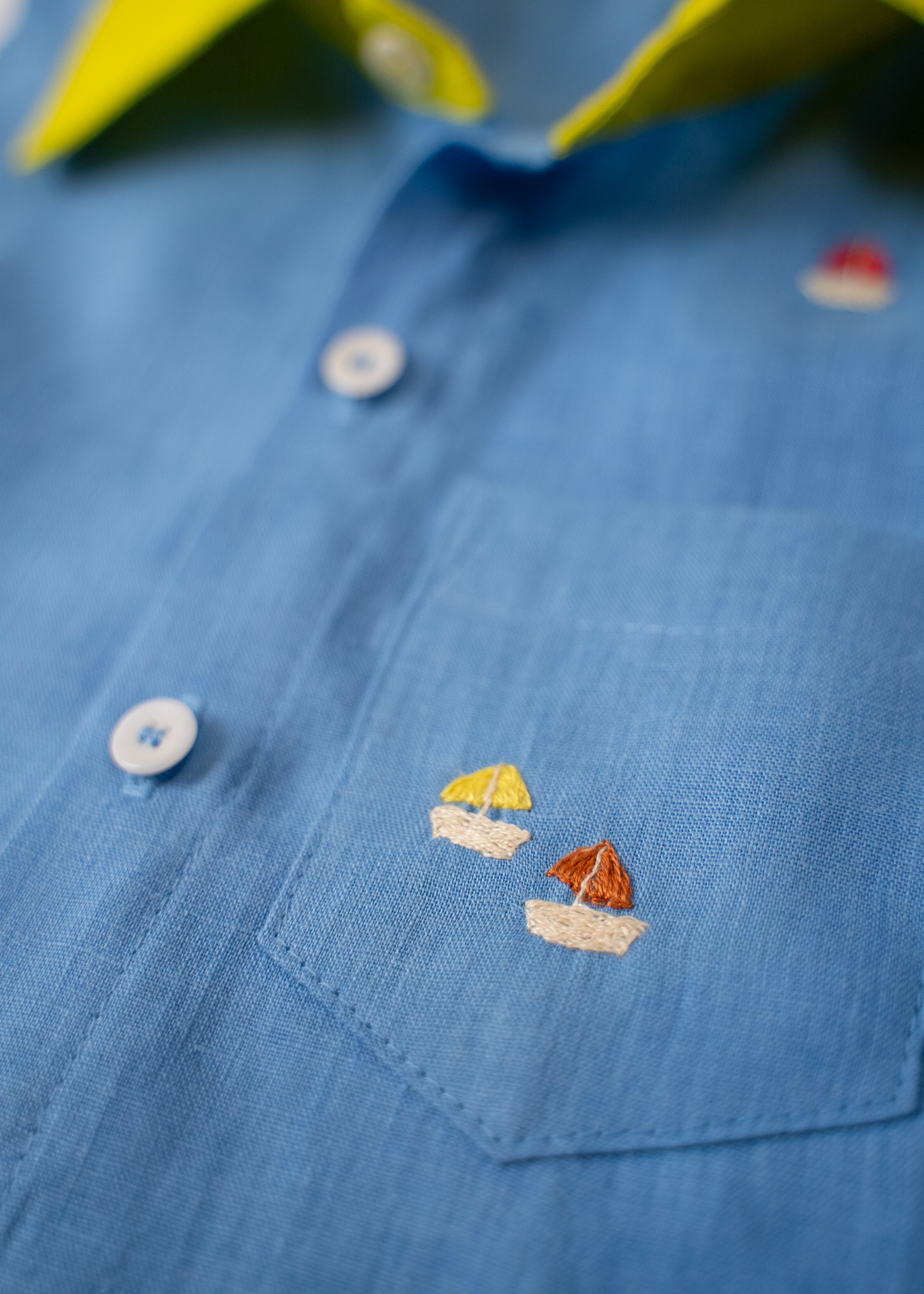 Detalhe do bolso com bordados à mão de barquinhos da camisa infantil em linho azul com gola amarelo lima.