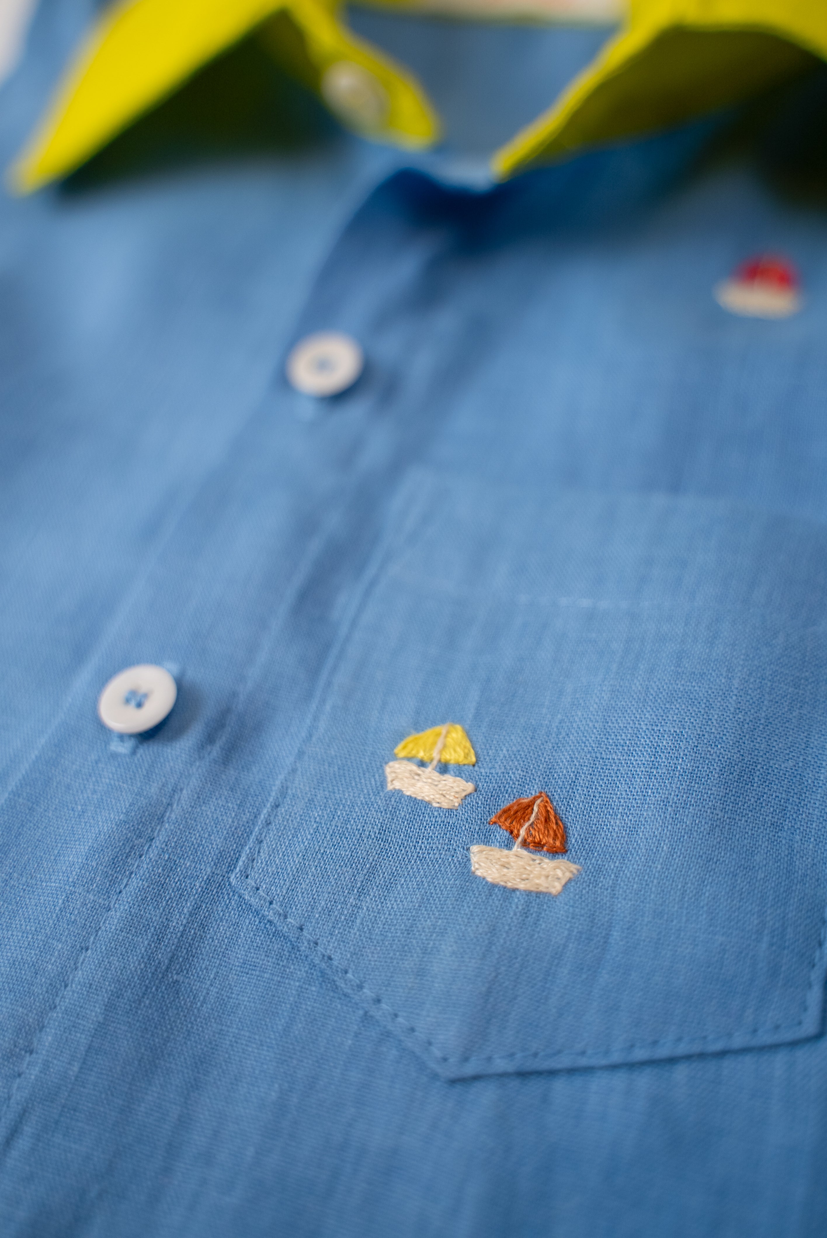 Detalhe do bolso com bordados à mão de barquinhos da camisa infantil em linho azul com gola amarelo lima.