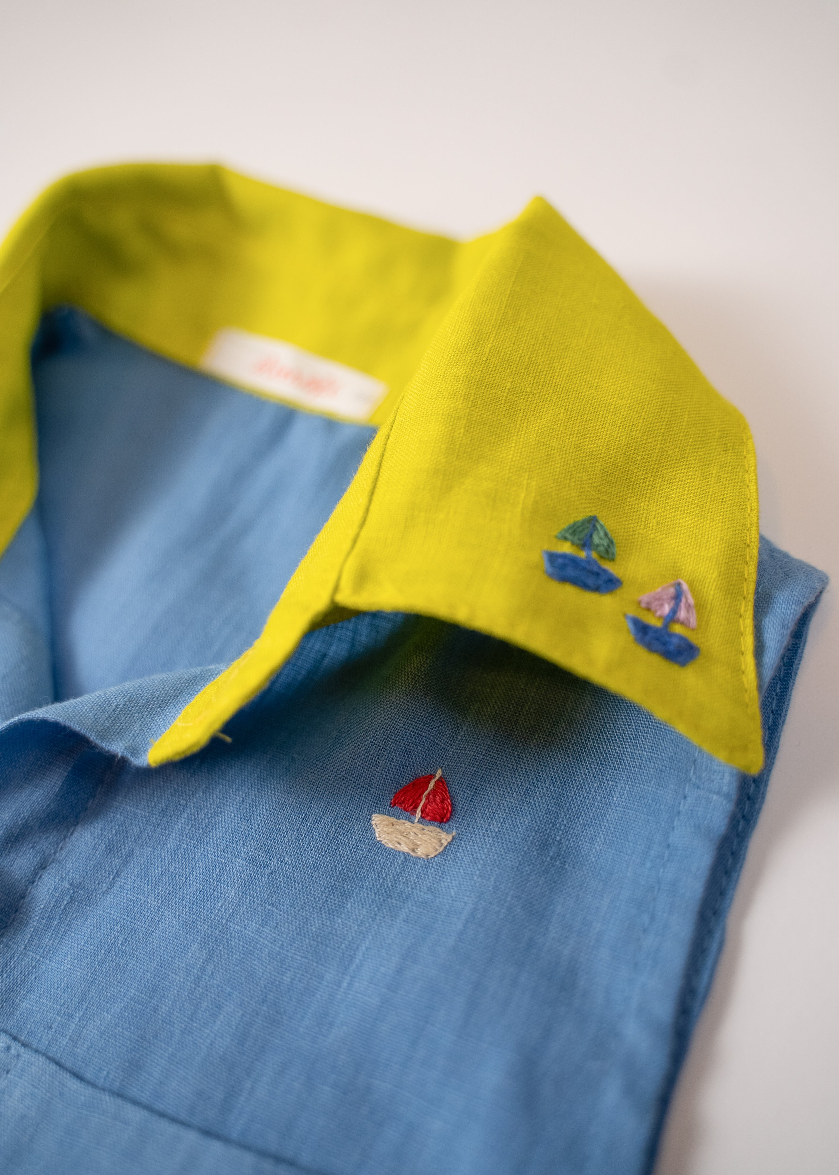 Detalhe da gola amarelo lima com bordados à mão de barquinhos da camisa infantil em linho azul celeste.