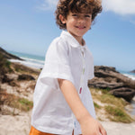 Menino usando camisa infantil em linho branco com bordados feitos à mão de peixes coloridos entre os botões.