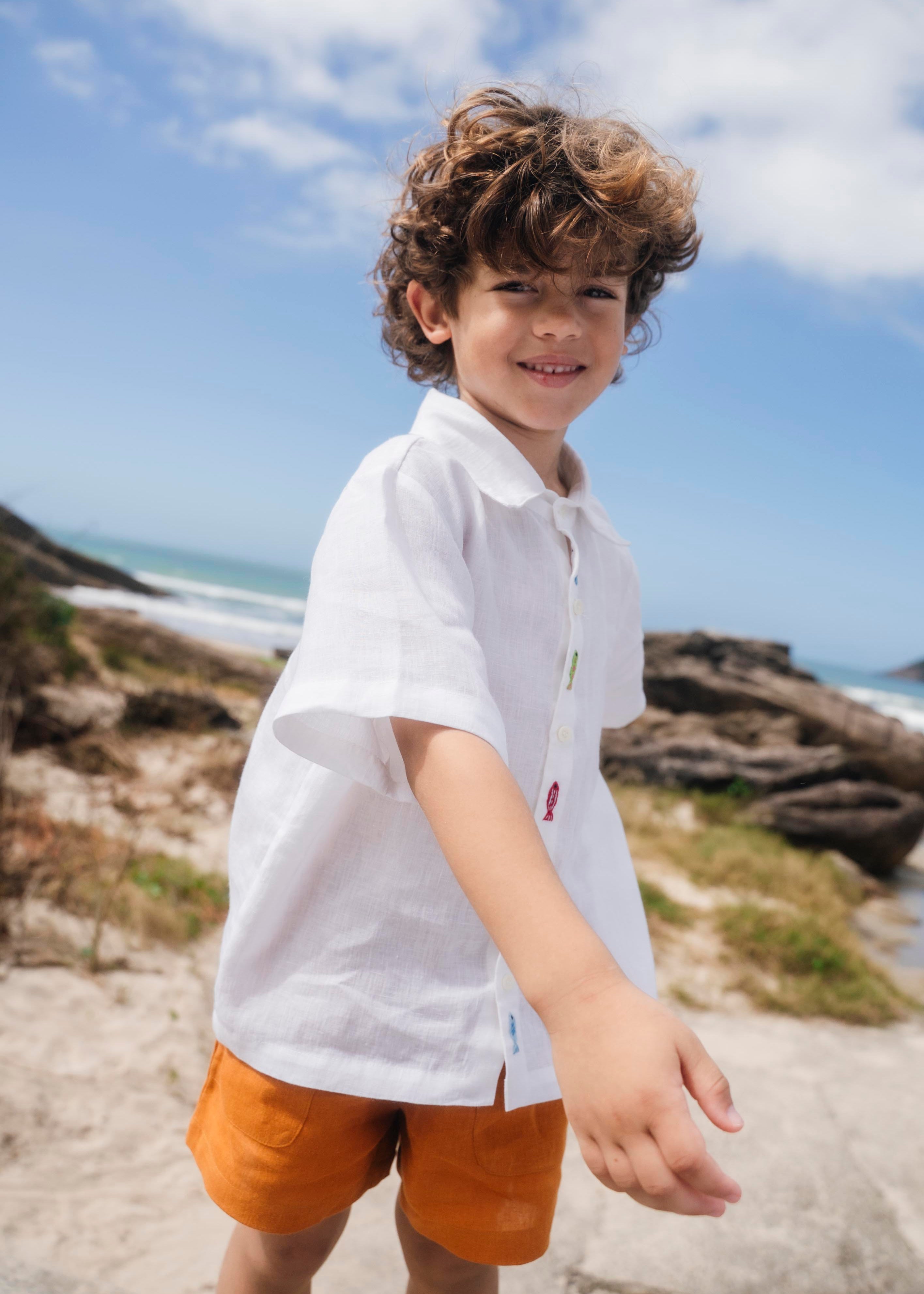 Menino usando camisa infantil em linho branco com bordados feitos à mão de peixes coloridos entre os botões.