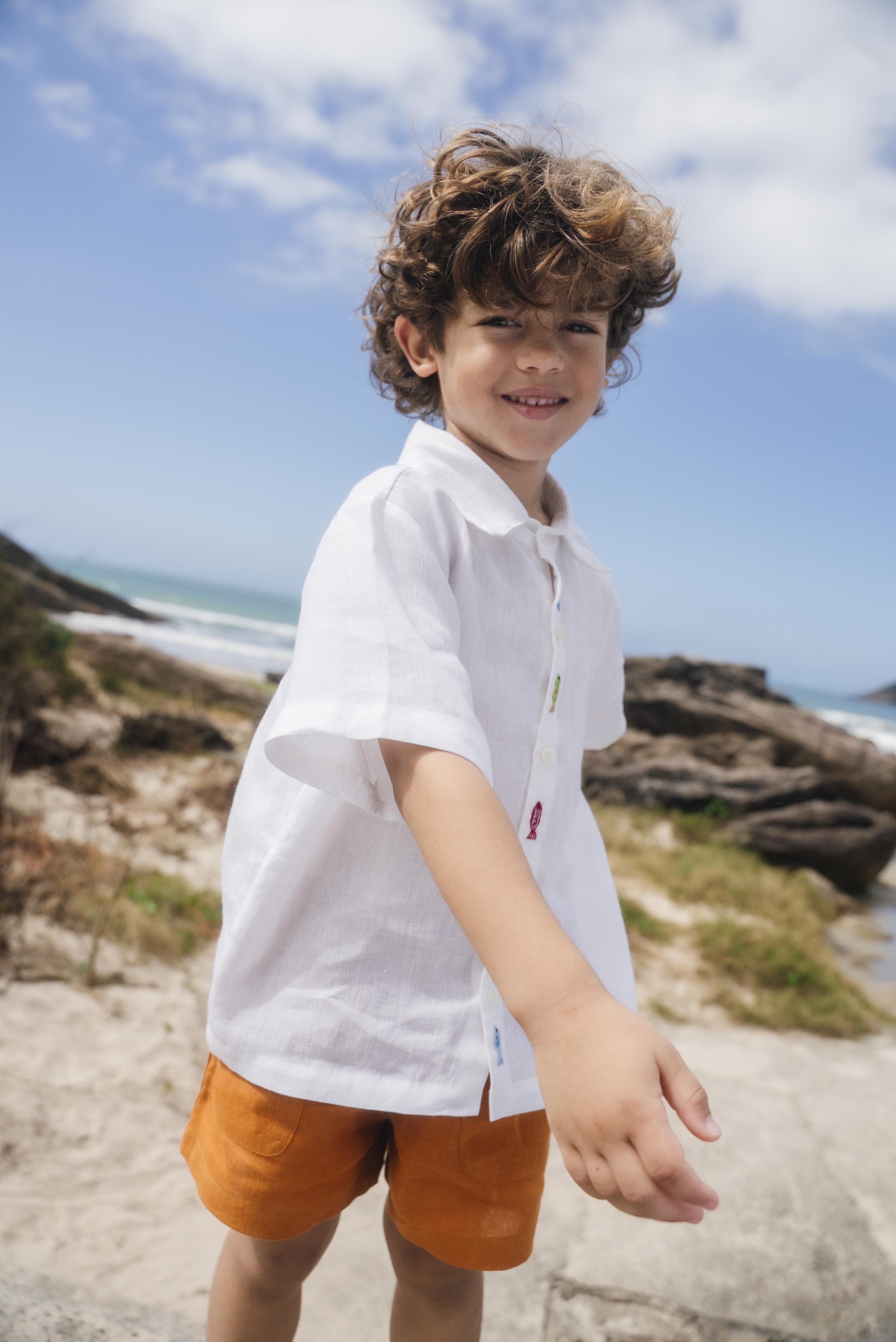 Menino usando camisa infantil em linho branco com bordados feitos à mão de peixes coloridos entre os botões.