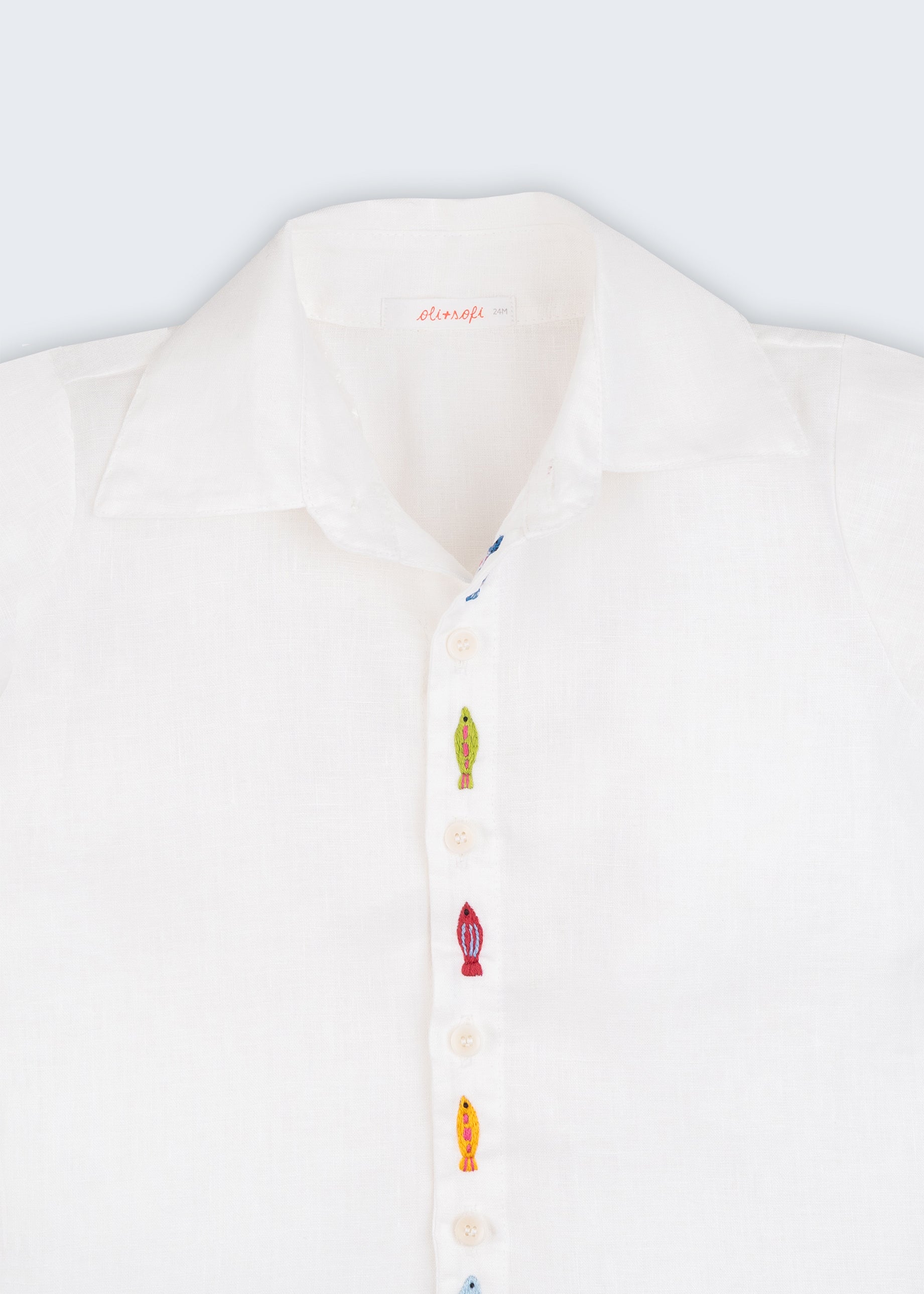 Camisa infantil em linho branco com bordados feitos à mão de peixes coloridos entre os botões, vista de frente em detalhe.