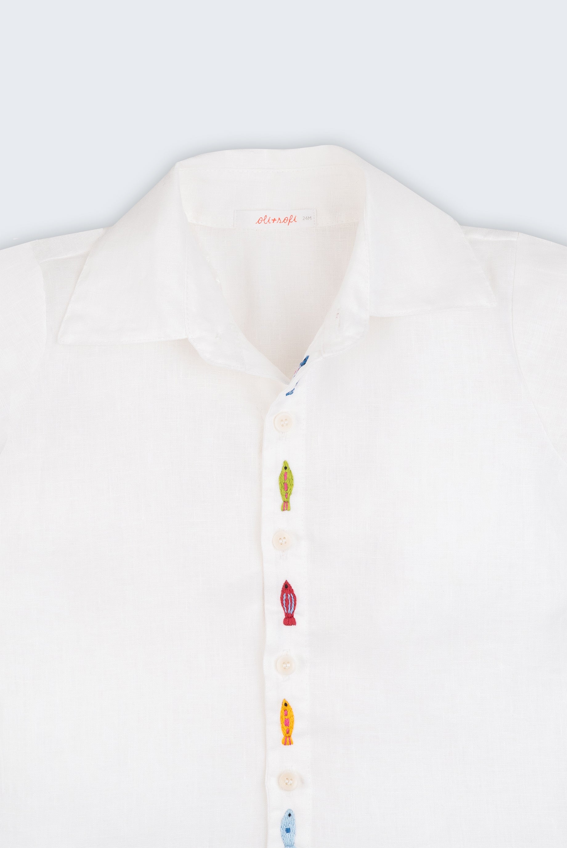Camisa infantil em linho branco com bordados feitos à mão de peixes coloridos entre os botões, vista de frente em detalhe.