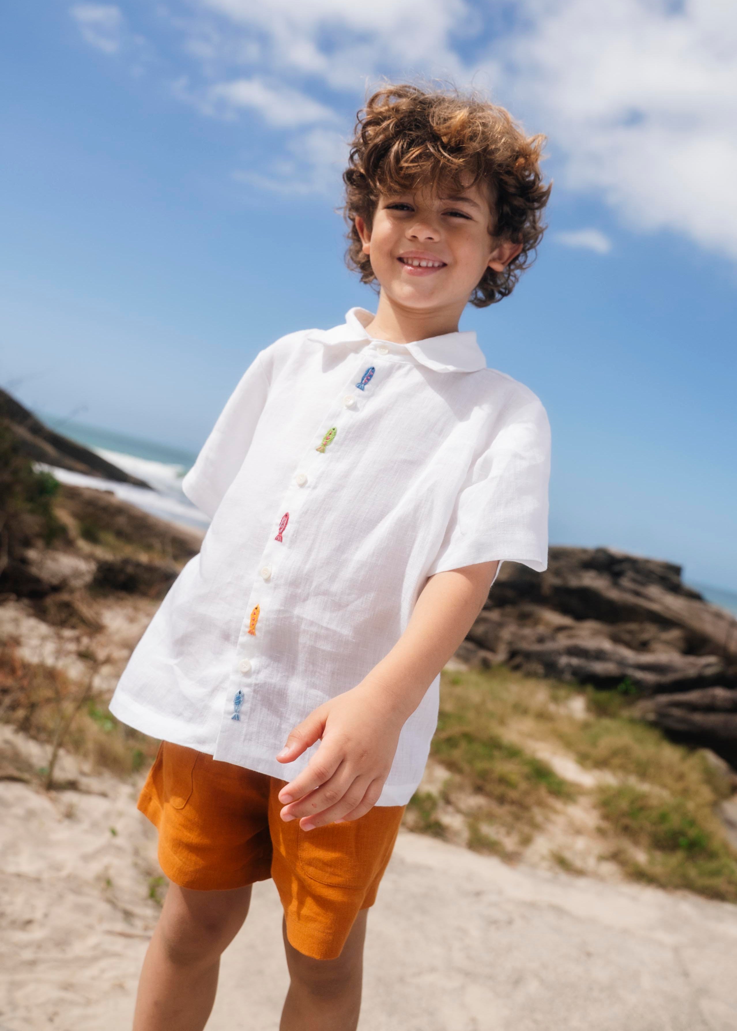 Menino com camisa infantil em linho branco com bordados feitos à mão de peixes coloridos entre os botões.