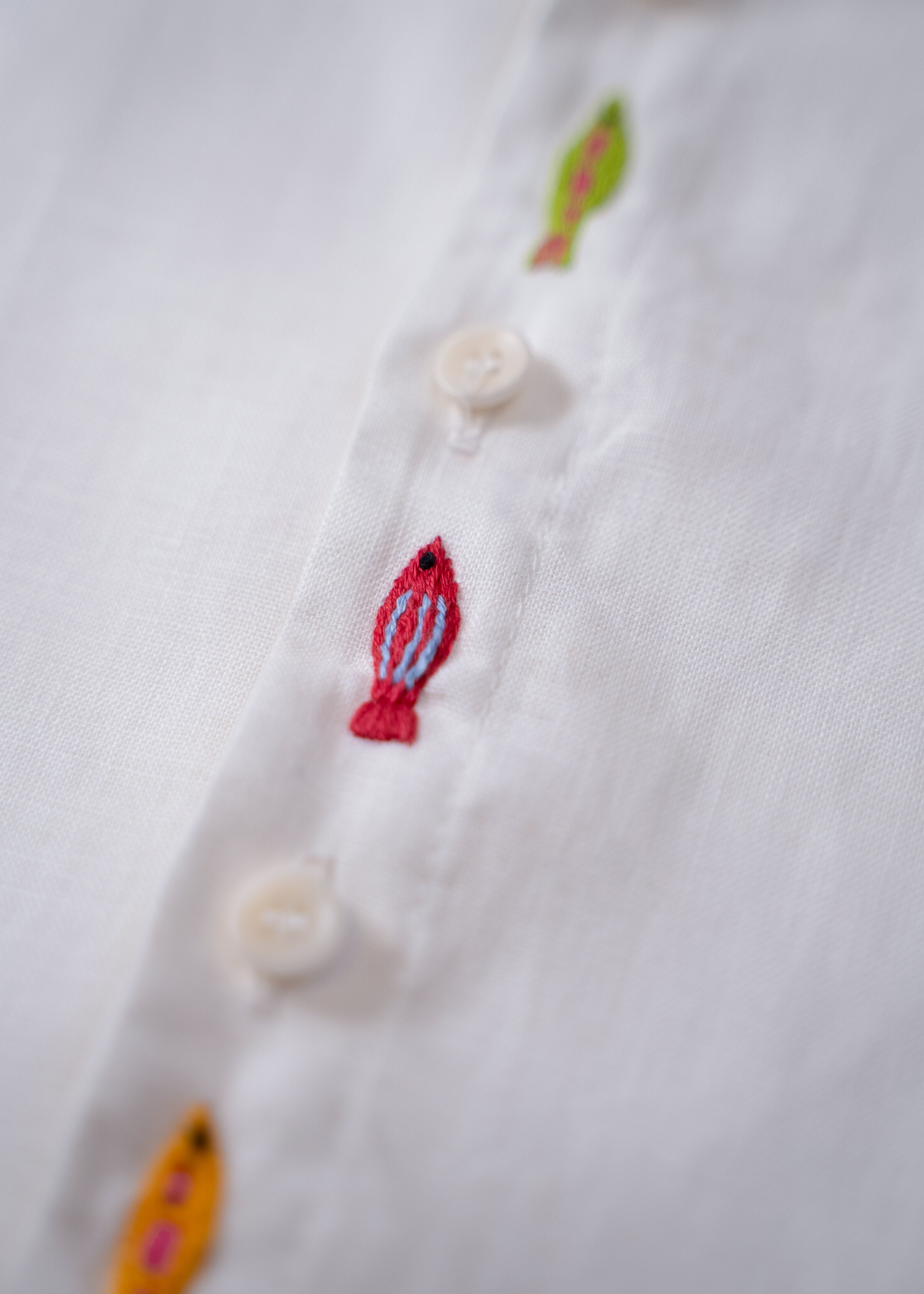 Detalhe do bordado à mão de peixe entre botões da camisa infantil em linho branco.