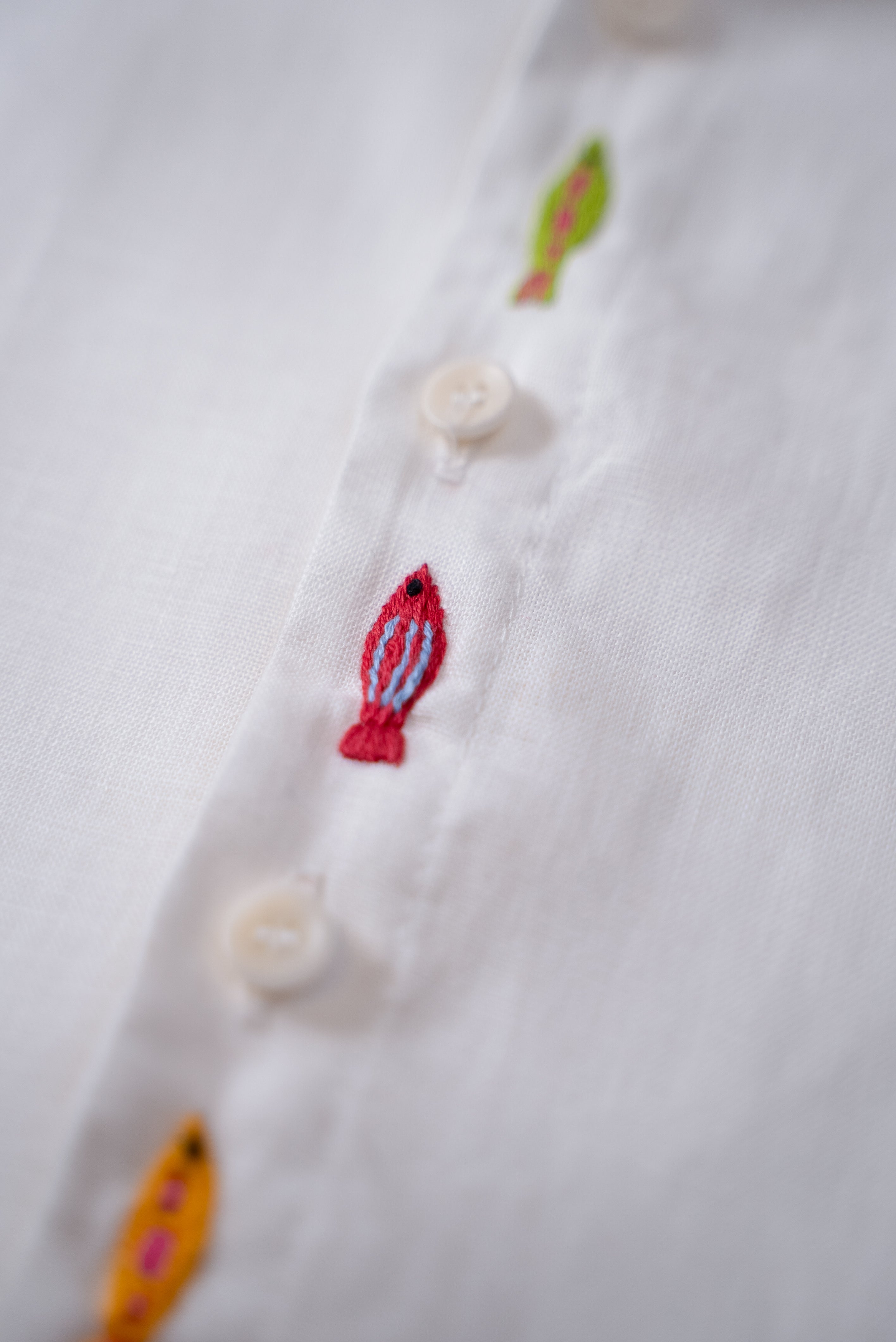 Detalhe do bordado à mão de peixe entre botões da camisa infantil em linho branco.