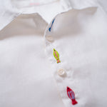 Detalhe do bordado à mão de peixe entre botões da camisa infantil unissex em linho branco.
