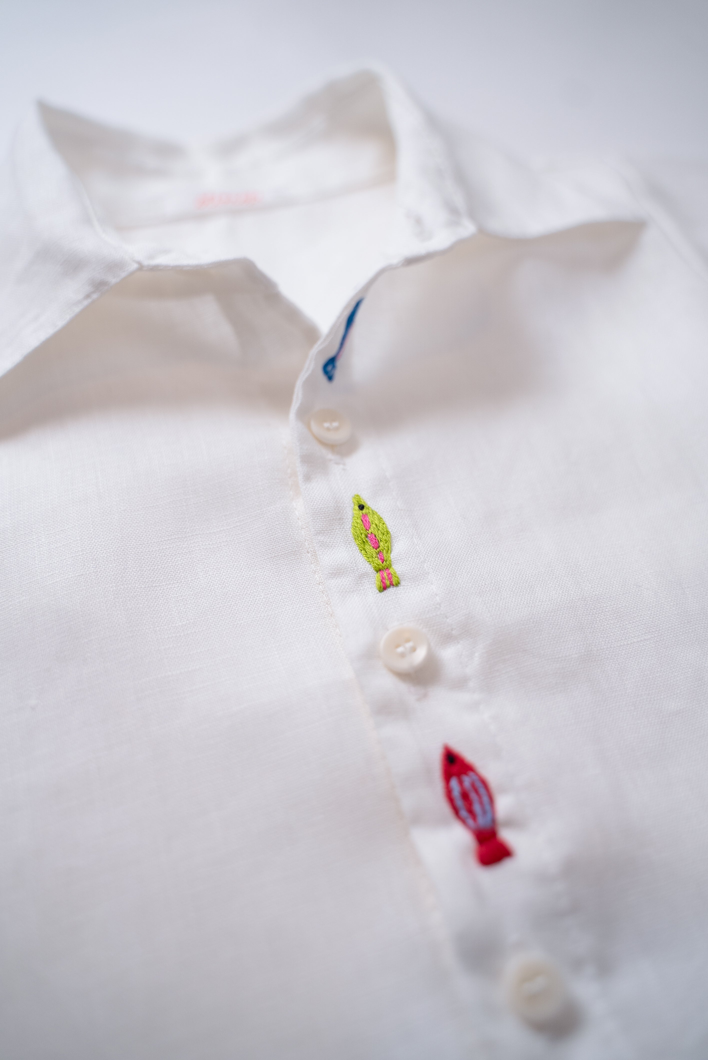 Detalhe do bordado à mão de peixe entre botões da camisa infantil unissex em linho branco.