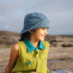 Menina sentada com camisa em linho verde com gola azul petróleo e bordado no peito de coqueiro feito à mão, com chapéu azul jeans na cabeça e shorts marrom.