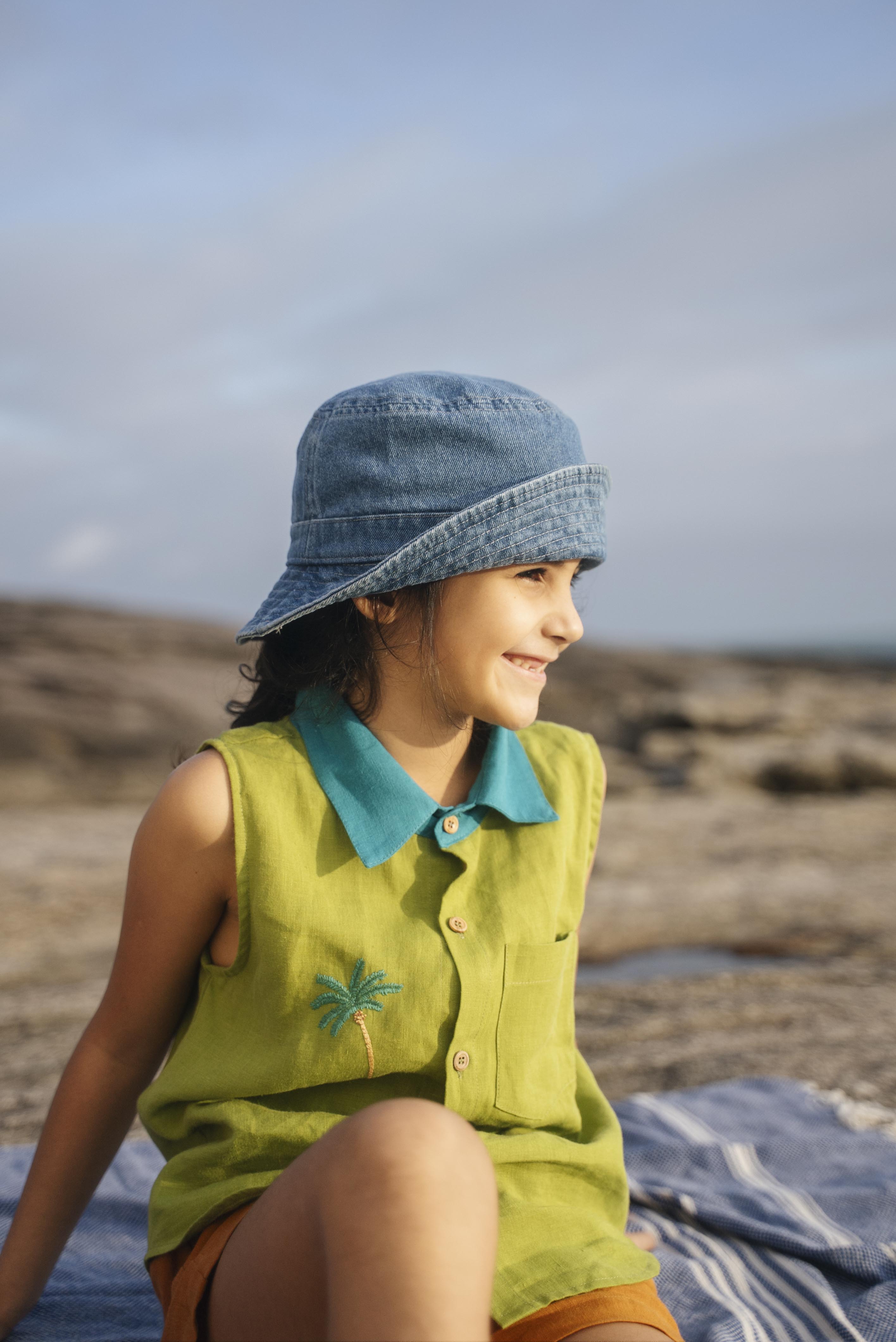 Menina sentada com camisa em linho verde com gola azul petróleo e bordado no peito de coqueiro feito à mão, com chapéu azul jeans na cabeça e shorts marrom.