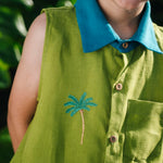 Menino usando camisa em linho verde com gola azul petróleo e bordado no peito de coqueiro e botões de madeira, visto em close.