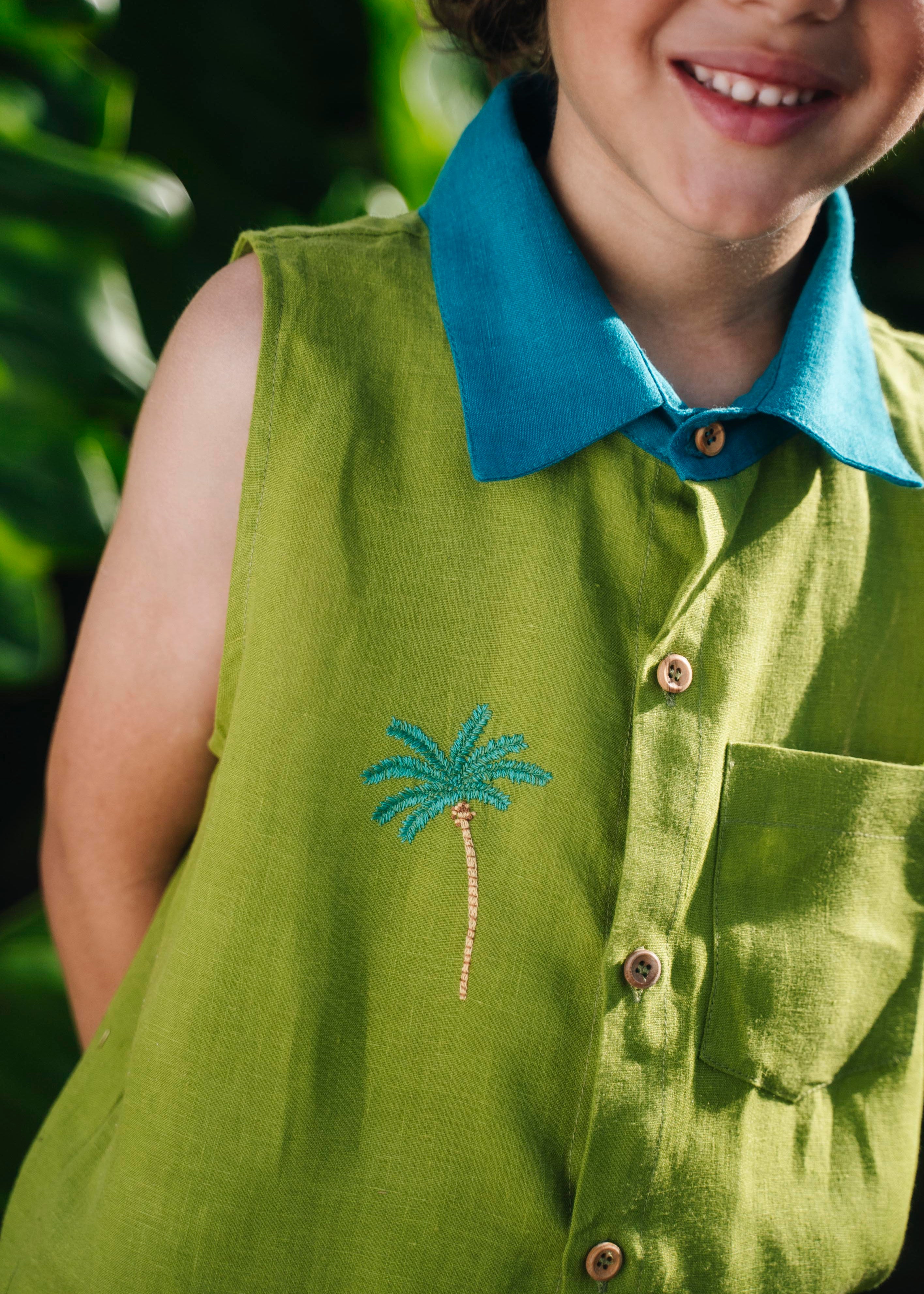 Menino usando camisa em linho verde com gola azul petróleo e bordado no peito de coqueiro e botões de madeira, visto em close.