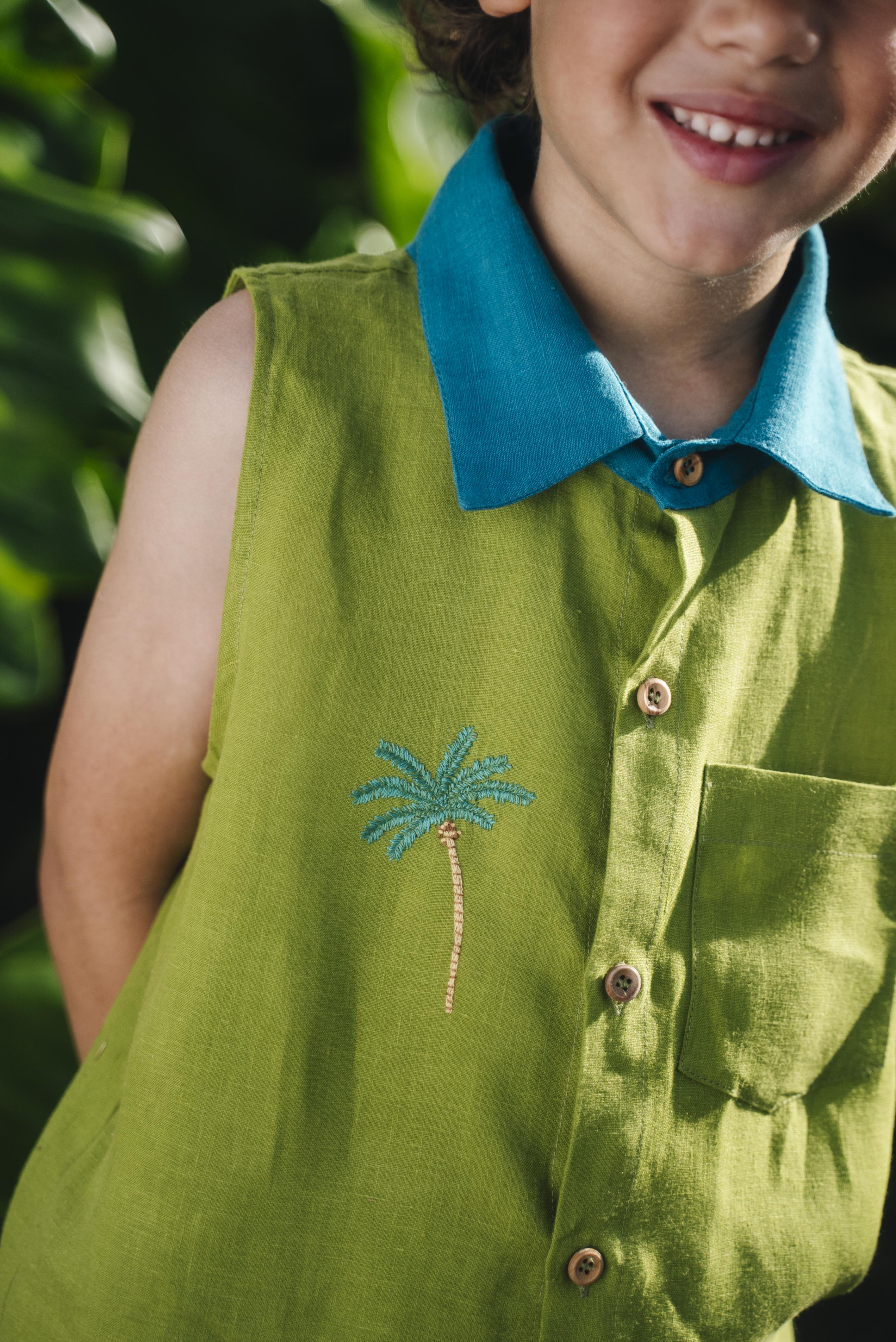 Menino usando camisa em linho verde com gola azul petróleo e bordado no peito de coqueiro e botões de madeira, visto em close.