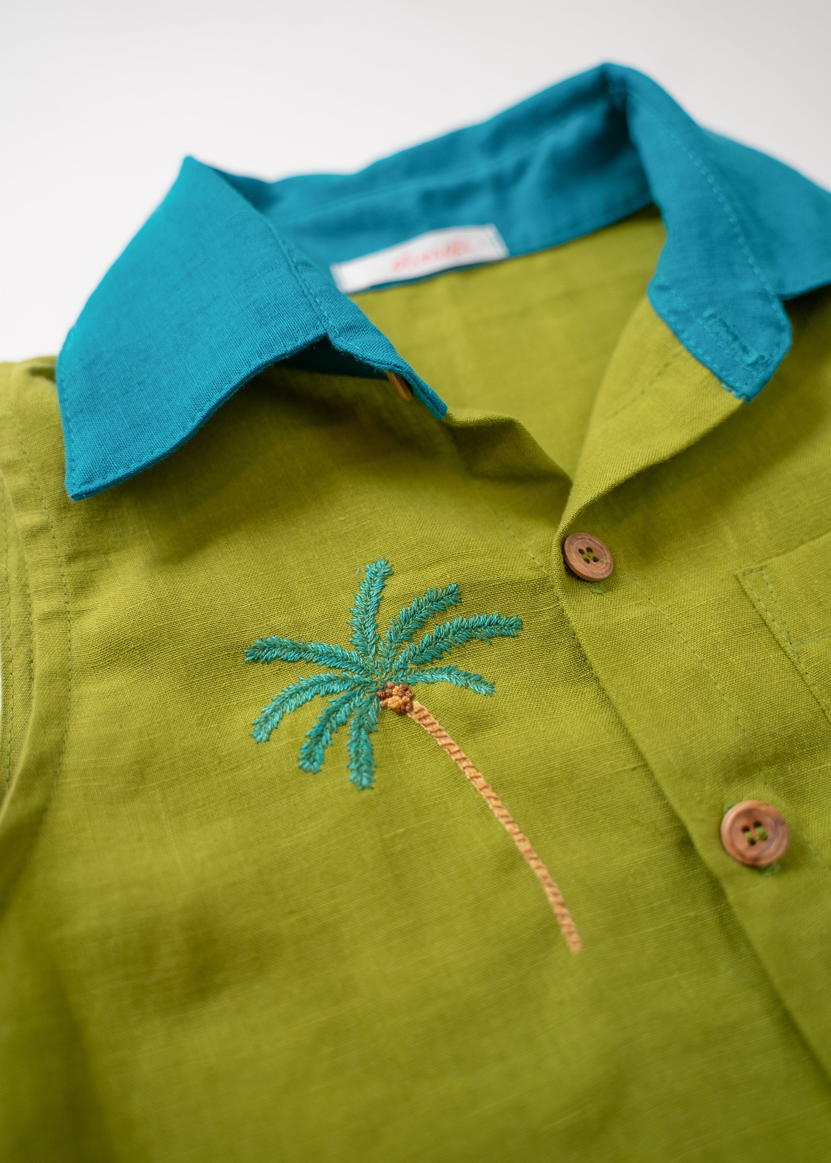 Detalhe do bordado à mão de coqueiro tropical no peito da camisa infantil em linho verde com gola azul petróleo.
