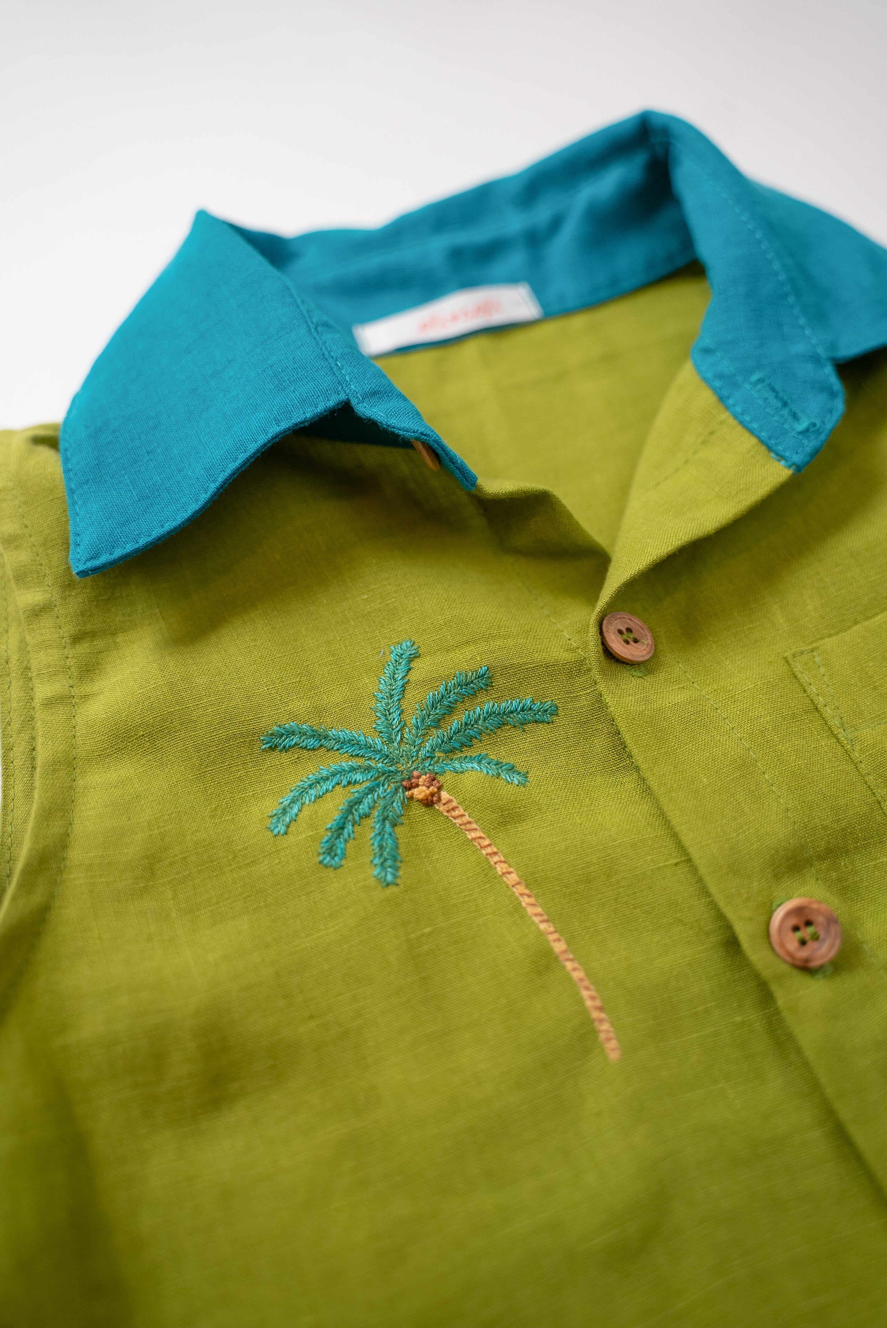 Detalhe do bordado à mão de coqueiro tropical no peito da camisa infantil em linho verde com gola azul petróleo.