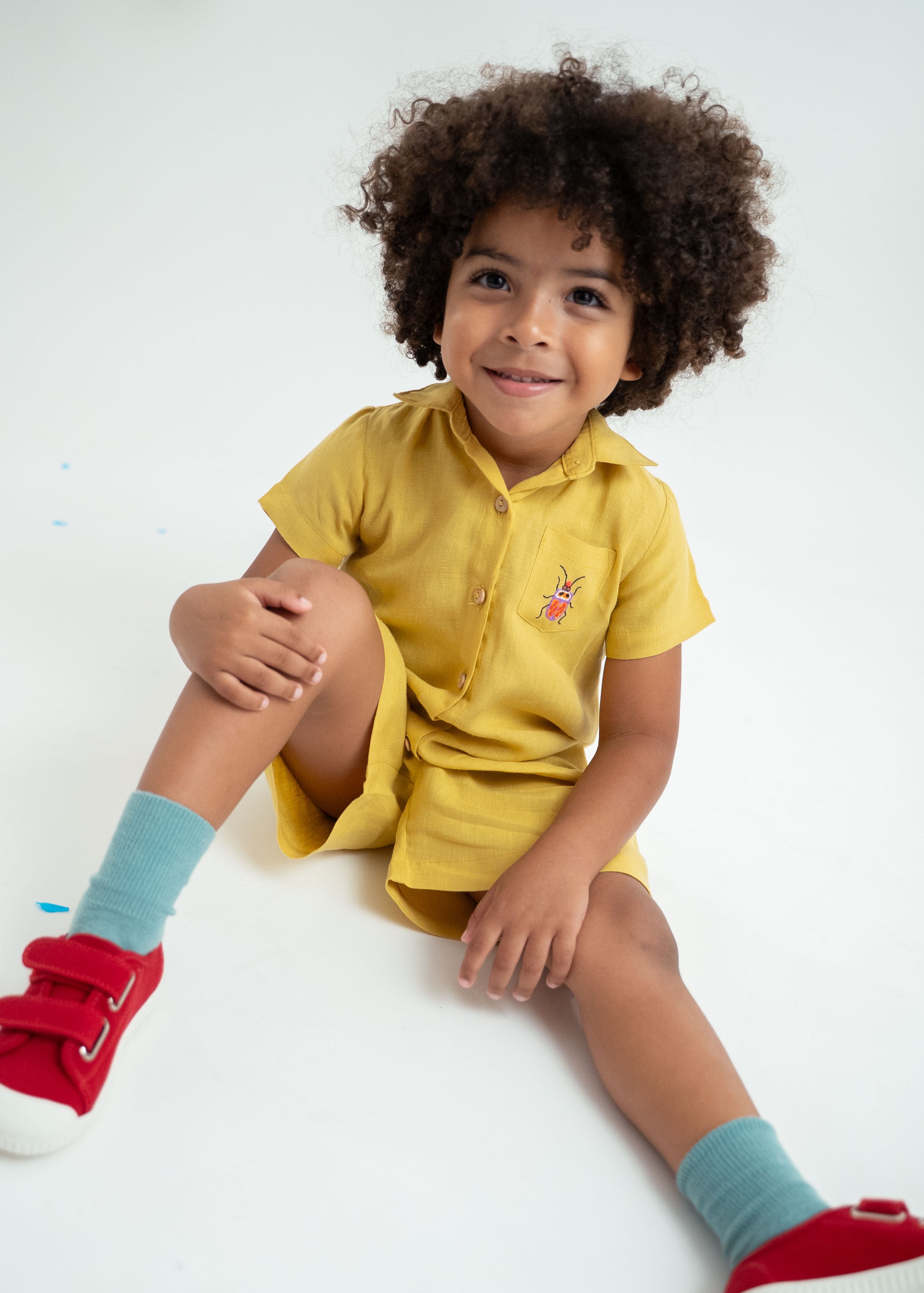Menino sentado vestindo macacão infantil unissex em linho amarelo com bordado de besouro no bolso.
