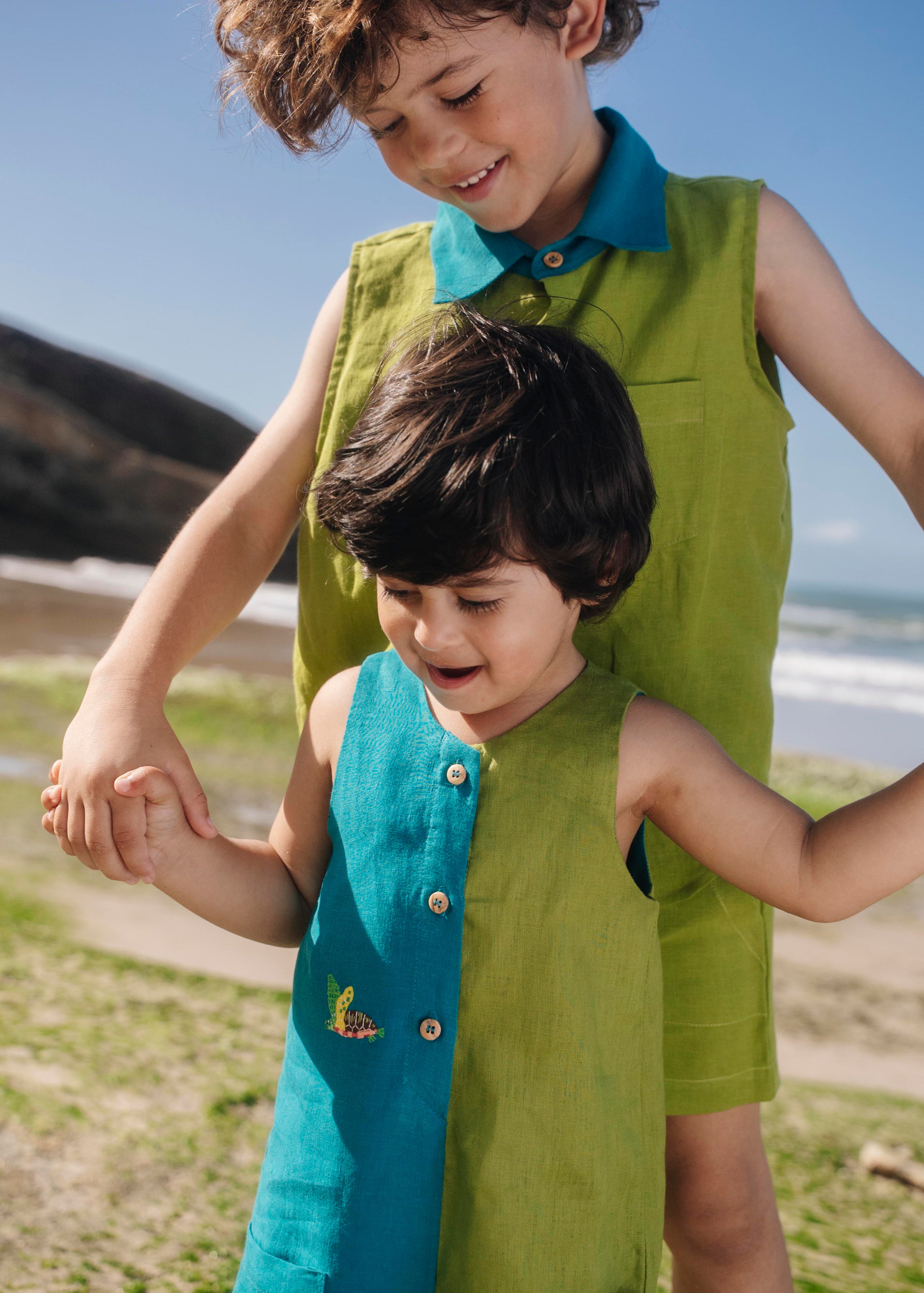 Menino usando macaquinho infantil em linho bicolor azul e verde com tartaruga bordada à mão, combinando com irmão.