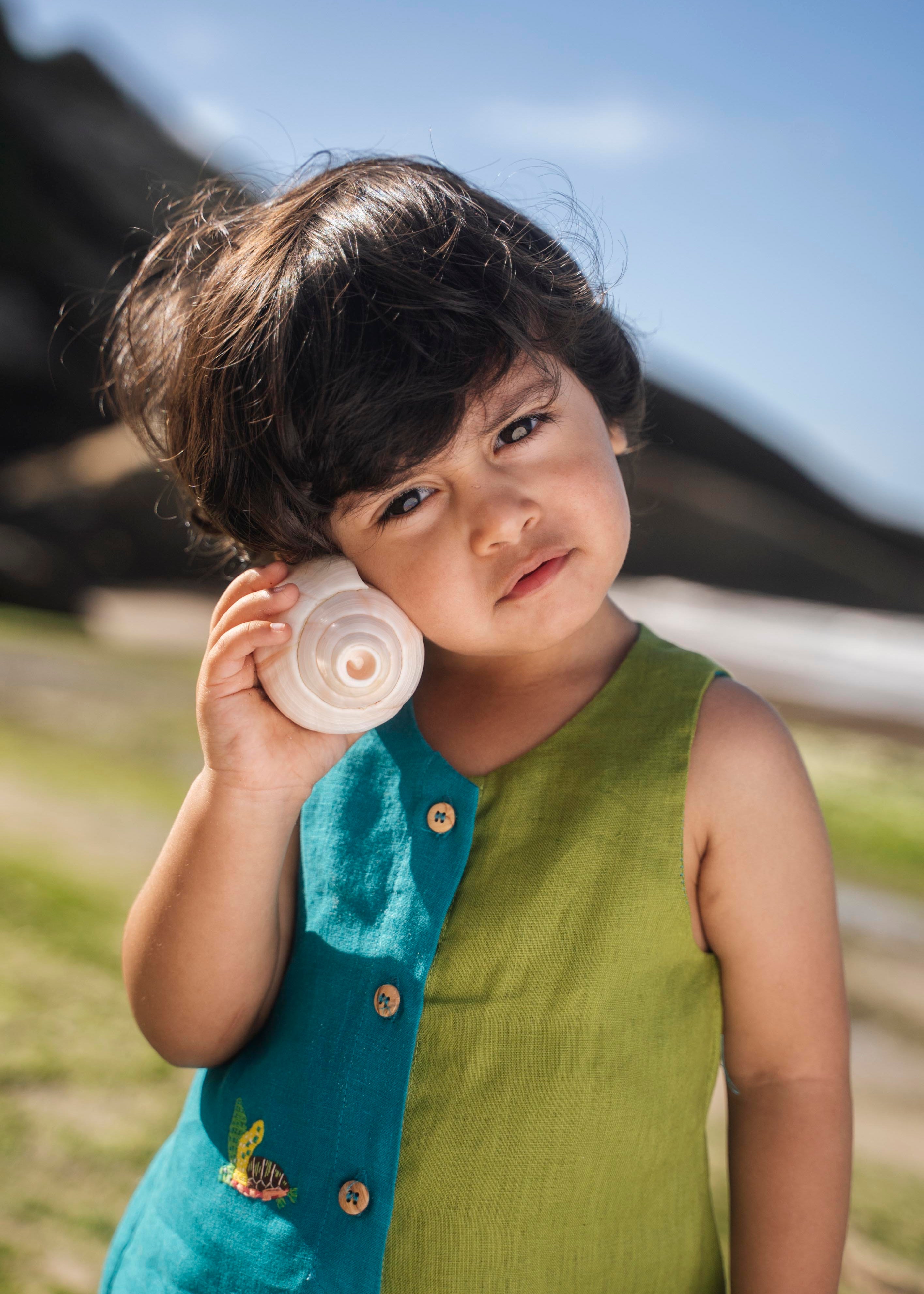 Menino usando macaquinho infantil em linho bicolor azul e verde com tartaruga bordada à mão, em close.