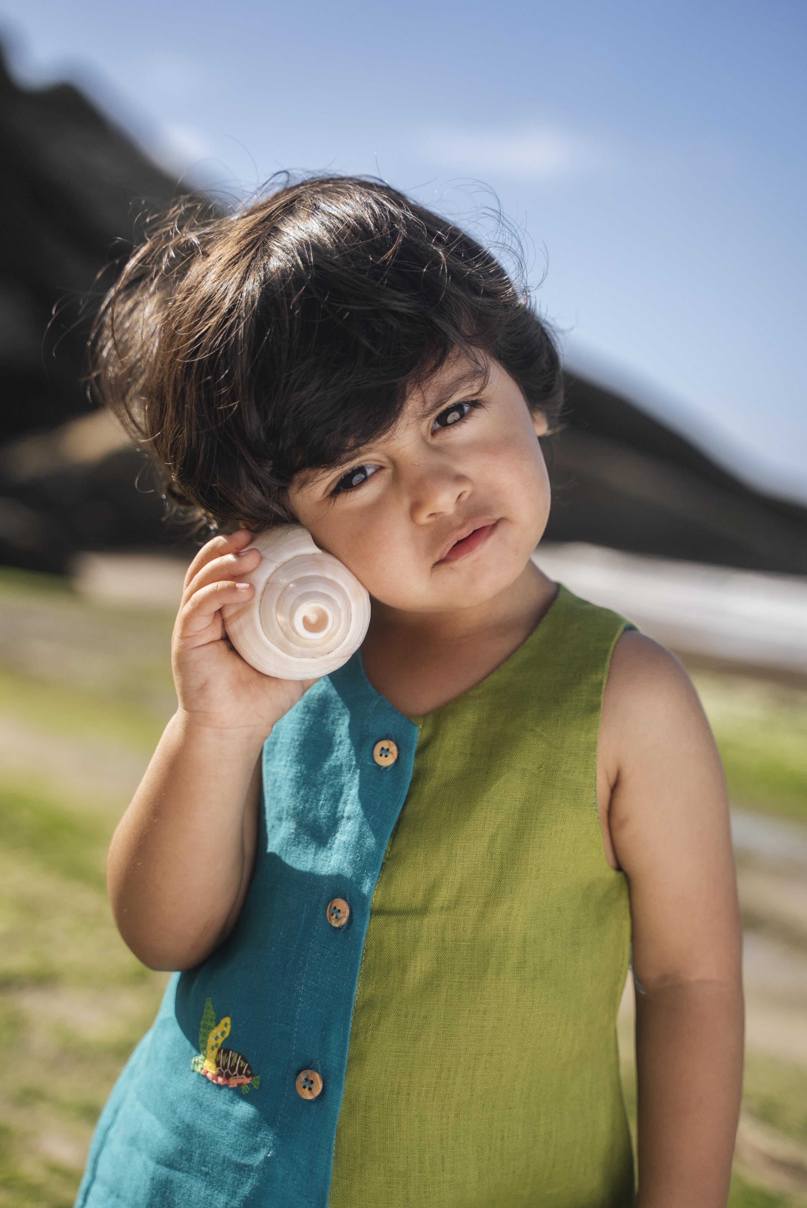 Menino usando macaquinho infantil em linho bicolor azul e verde com tartaruga bordada à mão, em close.